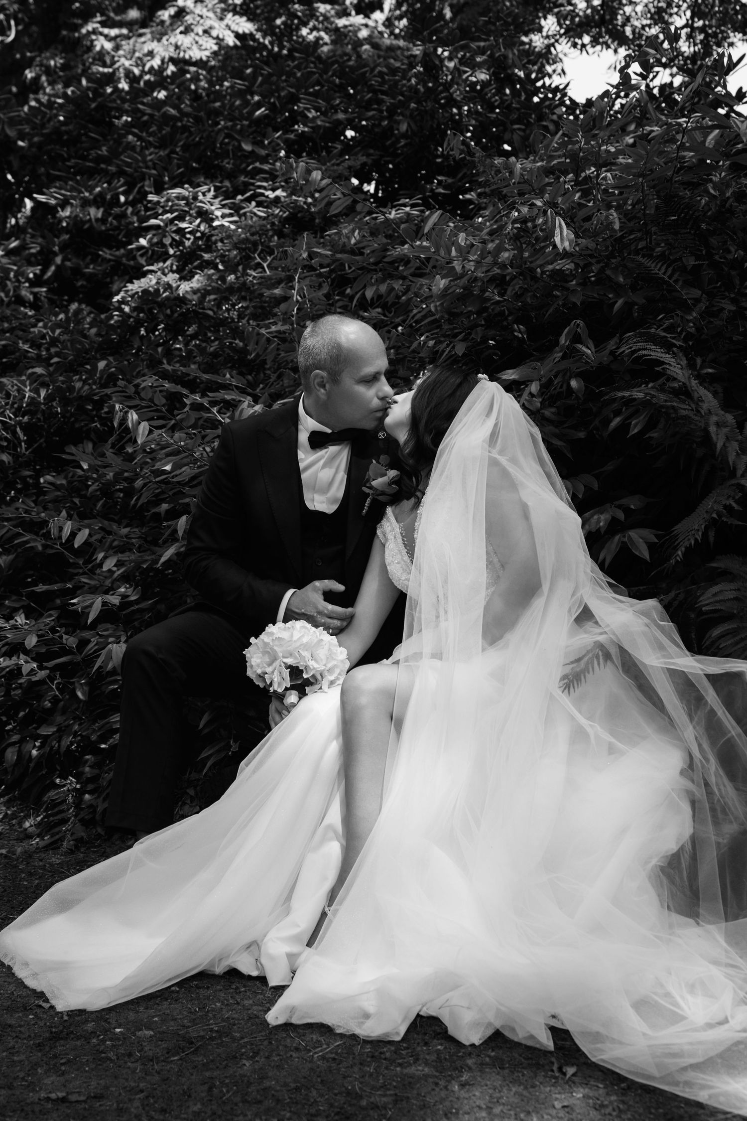 Black and white photo of a bride and groom sharing a kiss while seated outdoors among lush green foliage.