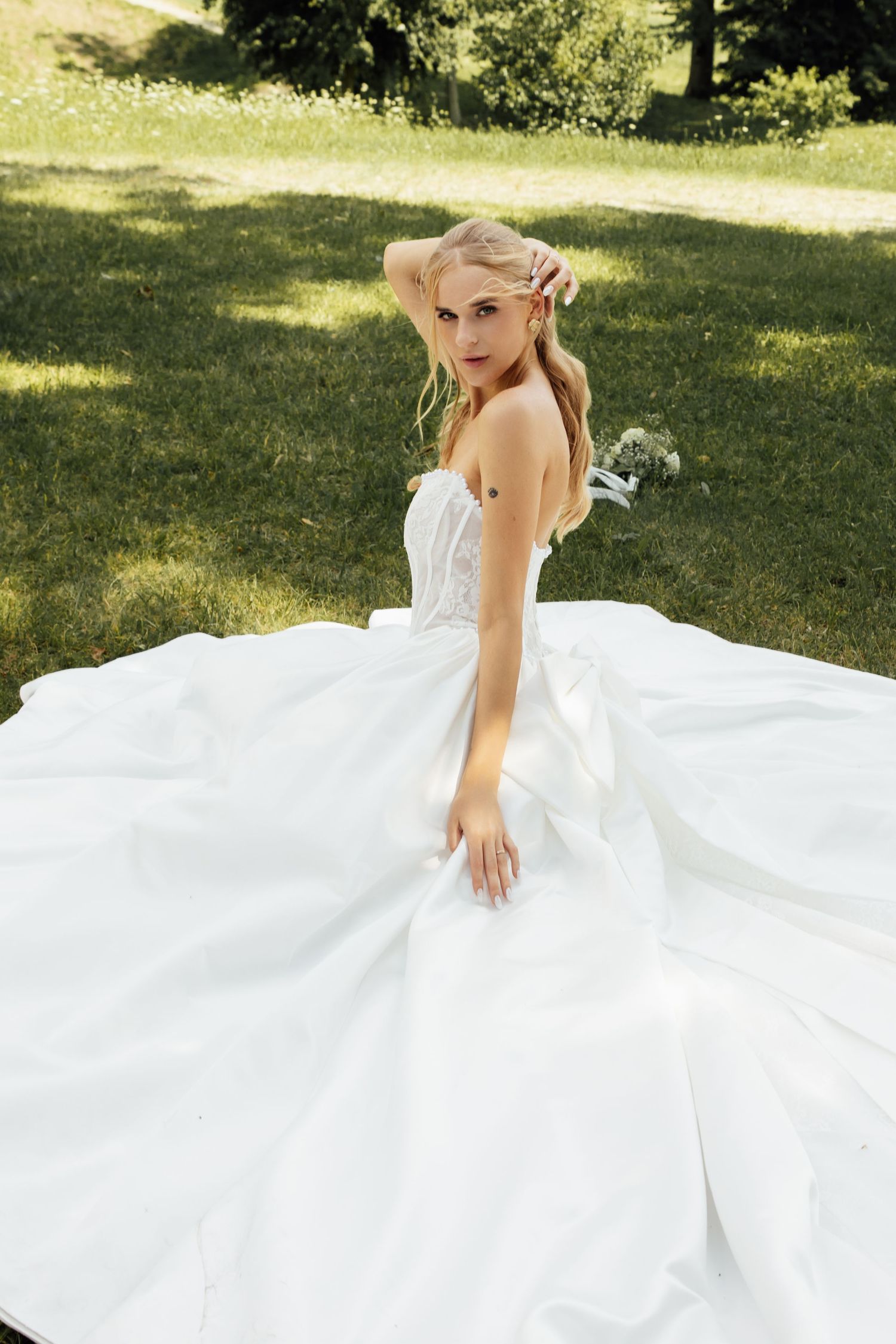 Elegant bride in a flowing white strapless ball gown sitting on green grass in a sunlit outdoor garden.