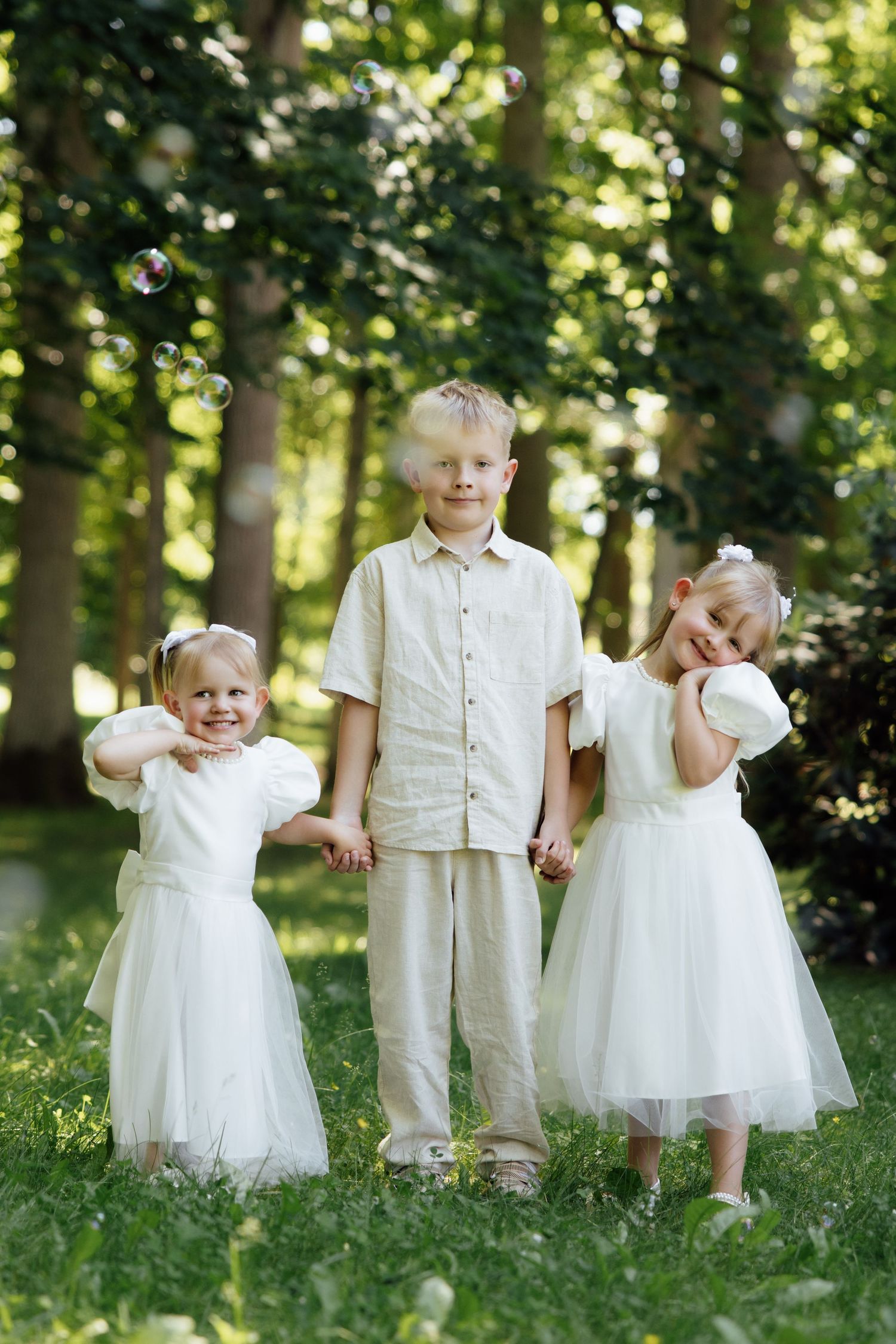 A boy in beige linen holds hands with two little girls in white tulle dresses among soap bubbles in a sunny park.