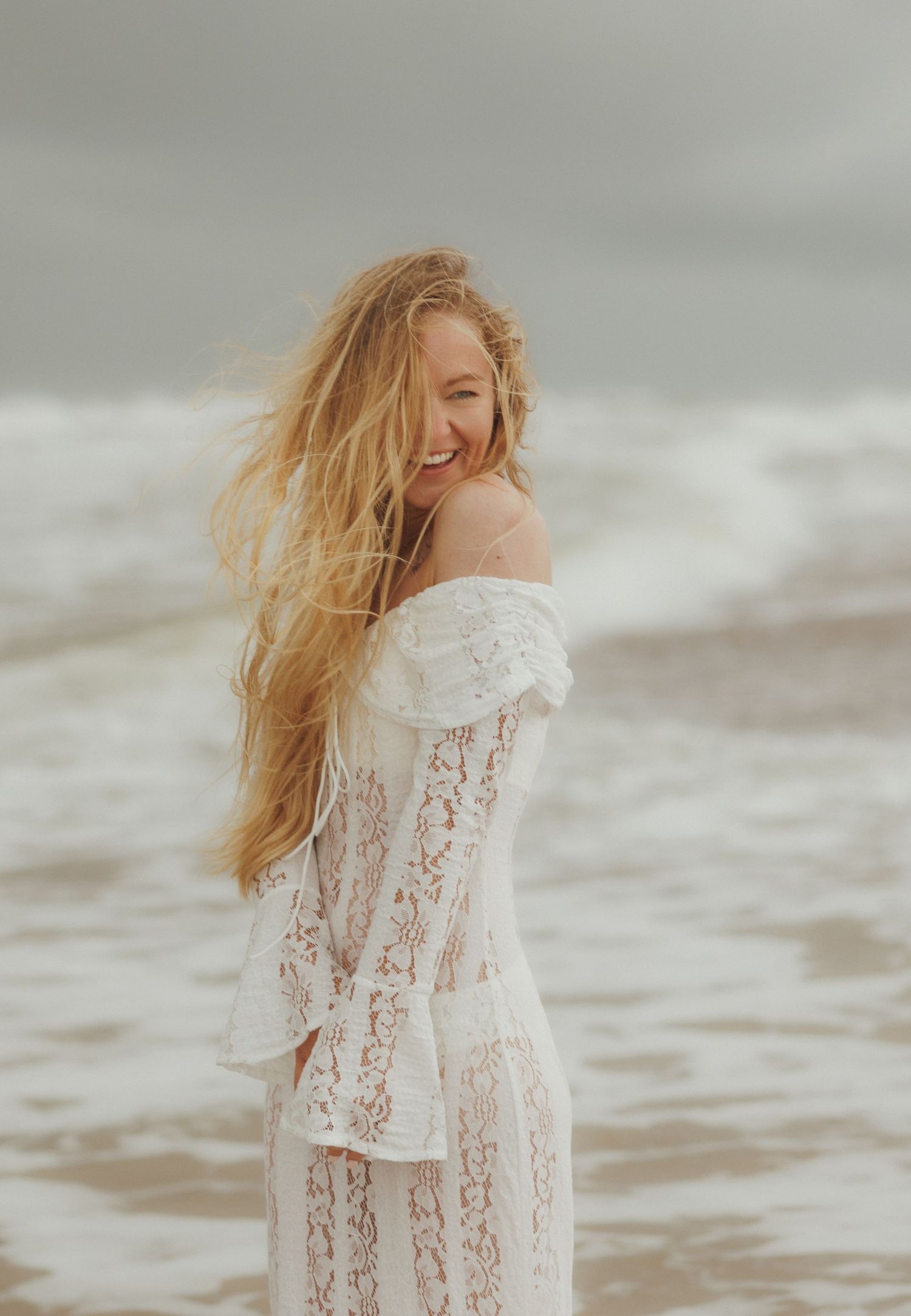 Smiling blonde woman in white lace off-shoulder dress standing on a windy beach with waves in the background.