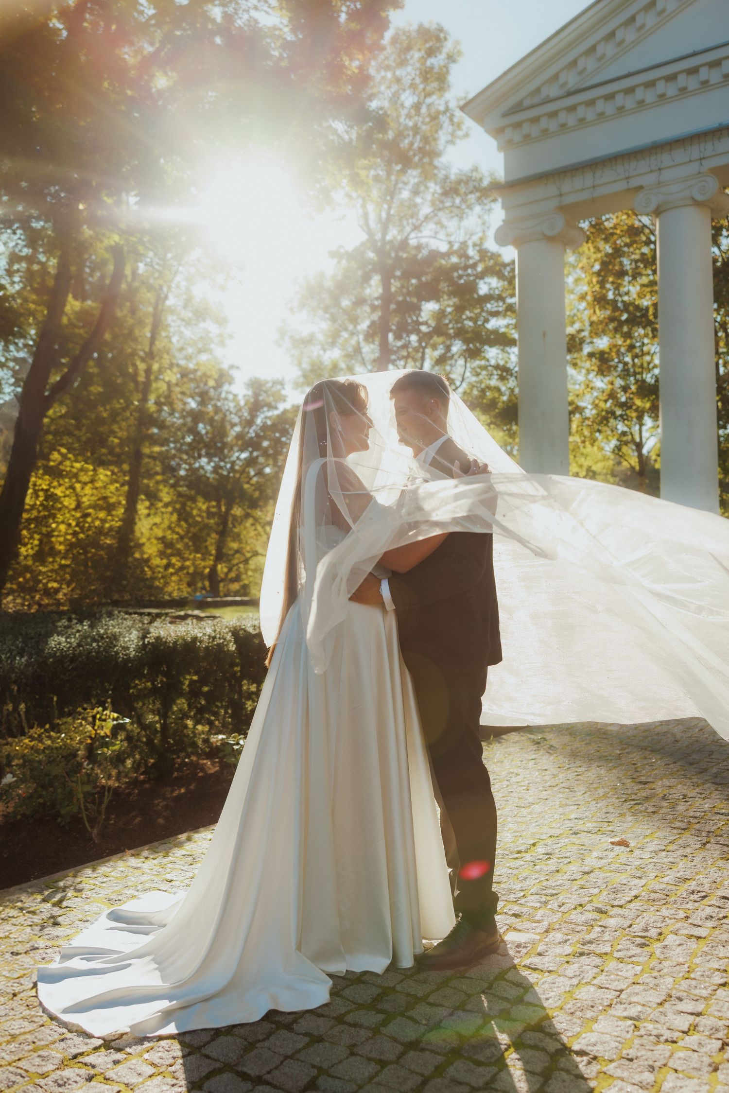 Romantic wedding moment captured under white columns with flowing dress and autumn leaves.