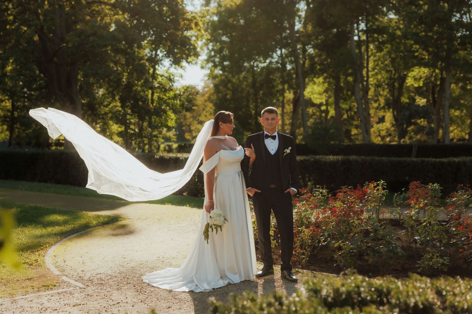 Wedding couple walks through a sunlit garden path with flowing dress and veil.