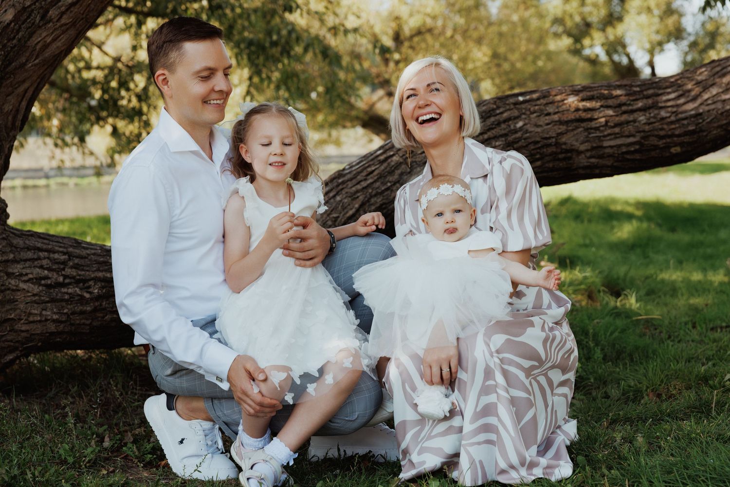 A joyful family wearing white outfits sits together on grass near a large tree trunk in a sunlit outdoor setting.