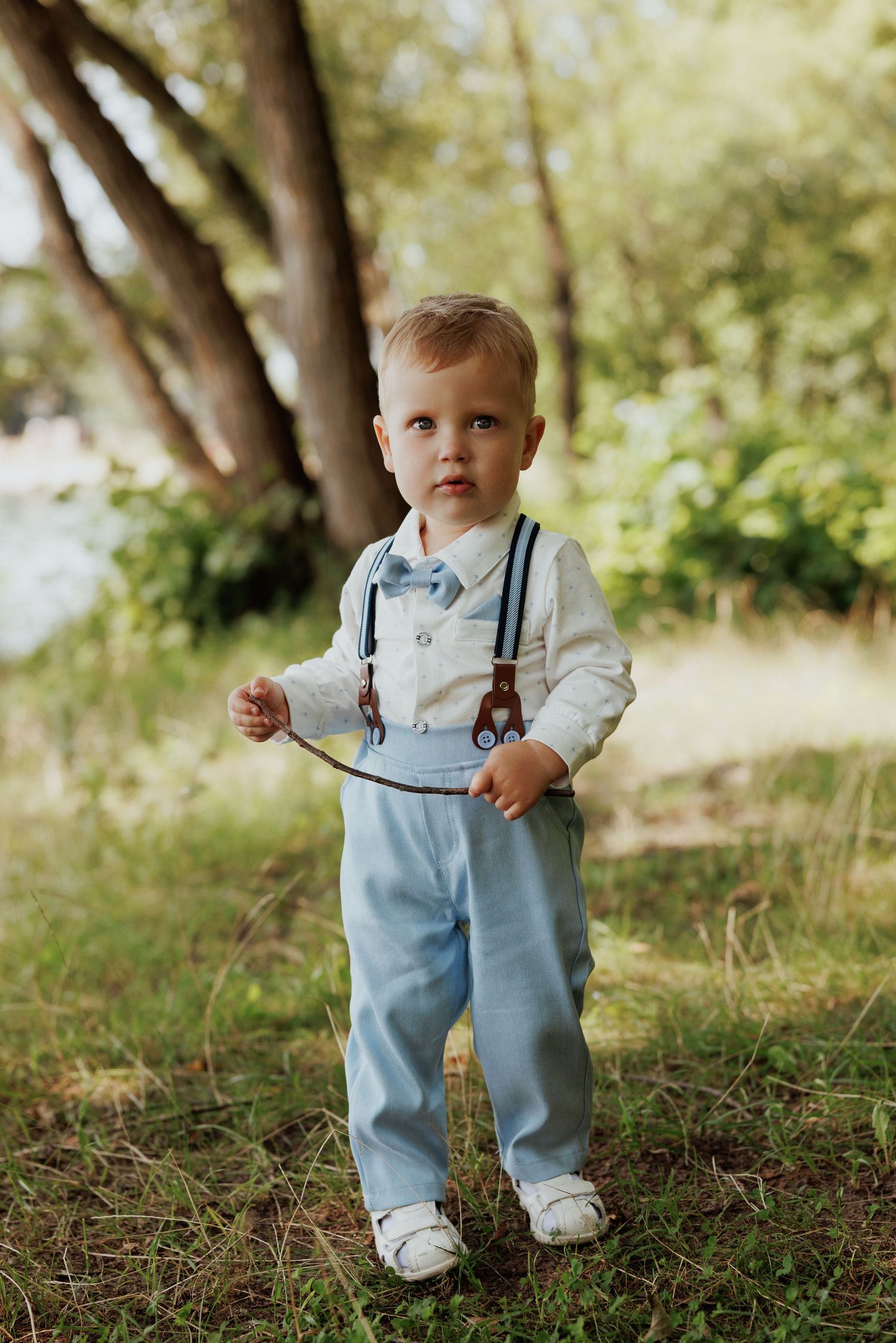 A small child wearing light blue overalls and white shirt walks through a sun-dappled wooded area in summer.