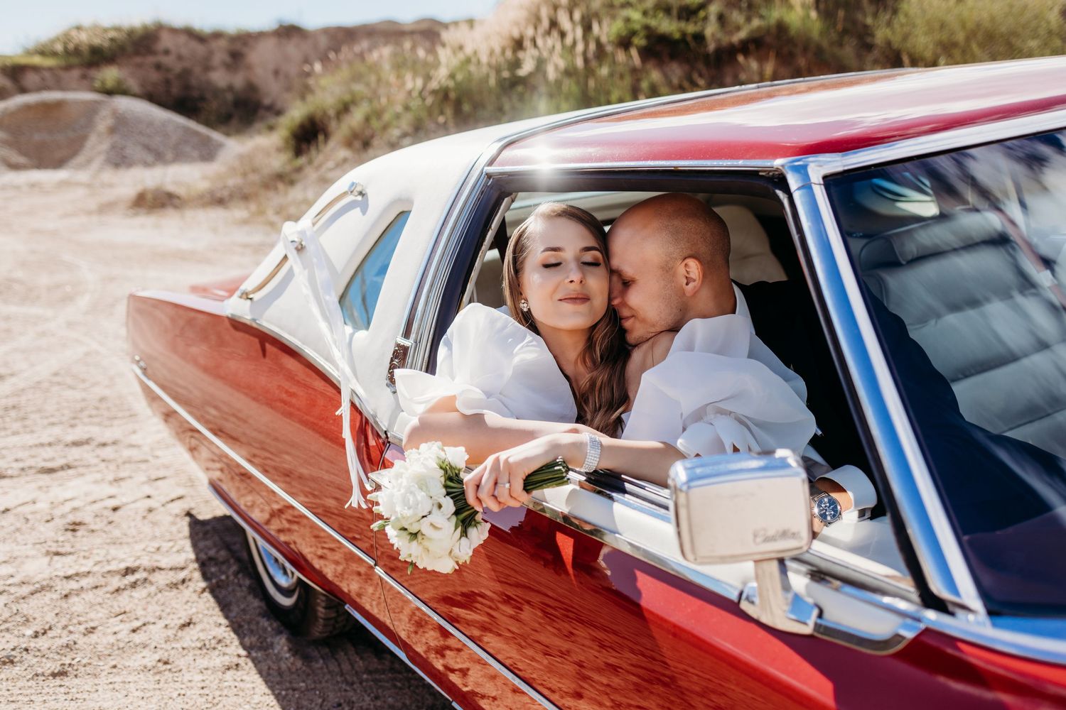 Newlyweds share intimate moments in classic red vintage car during wedding photoshoot.