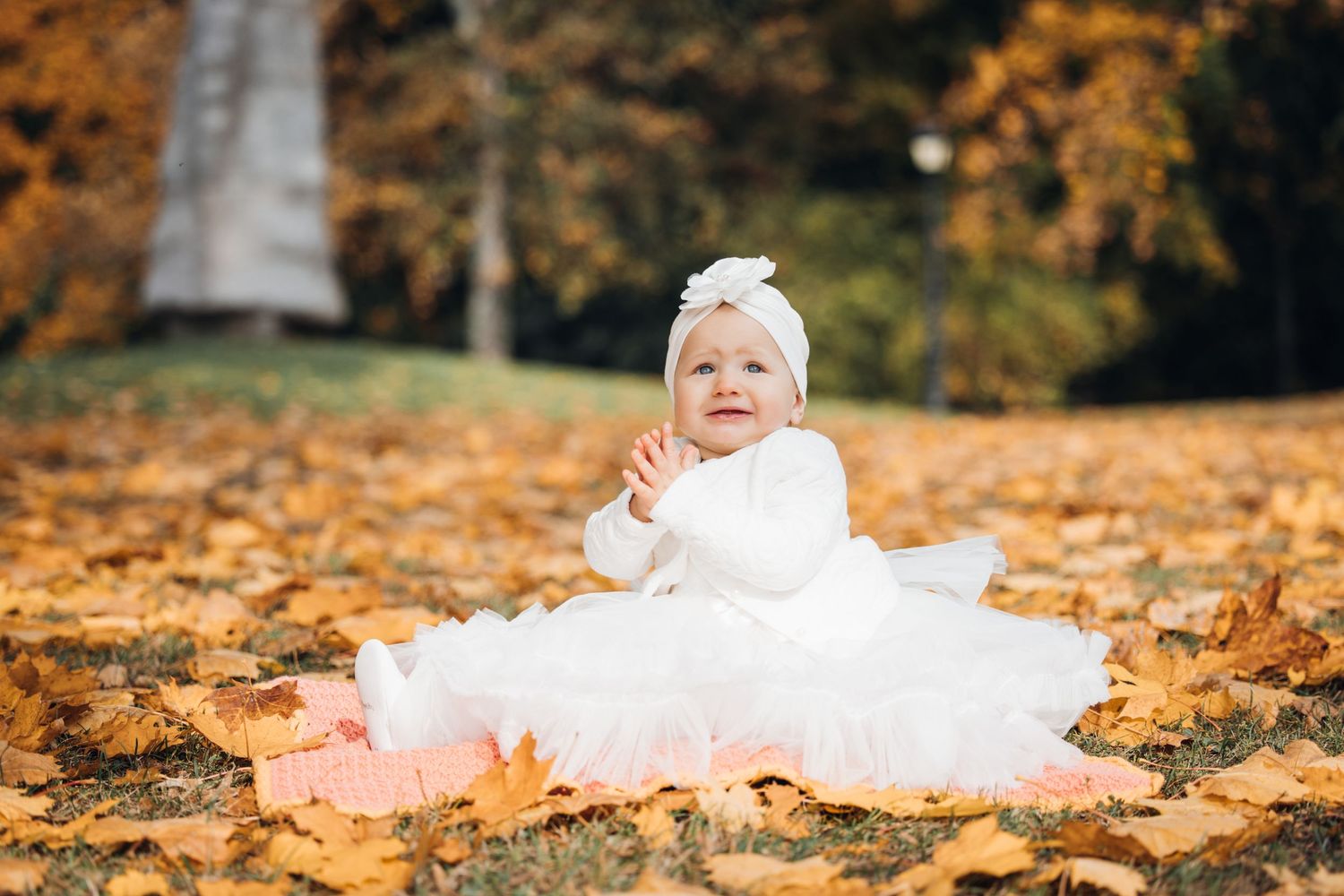 A baby in white lace dress lies on orange autumn leaves during an outdoor photo session.