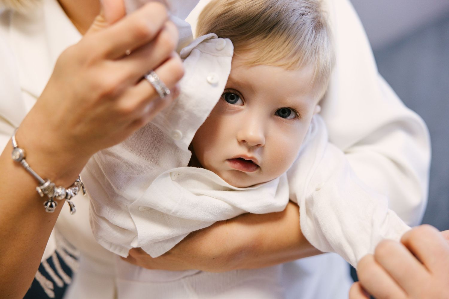 A baby in white clothing reaches up with their hand while being held against a soft gray background.