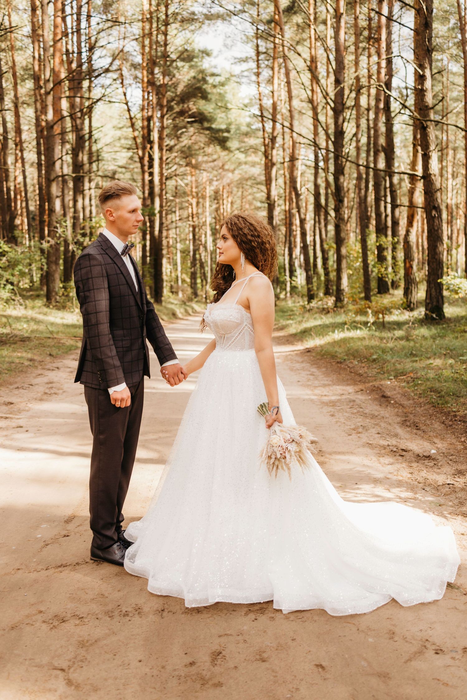 Wedding couple poses together on a sunlit forest path surrounded by tall pine trees.