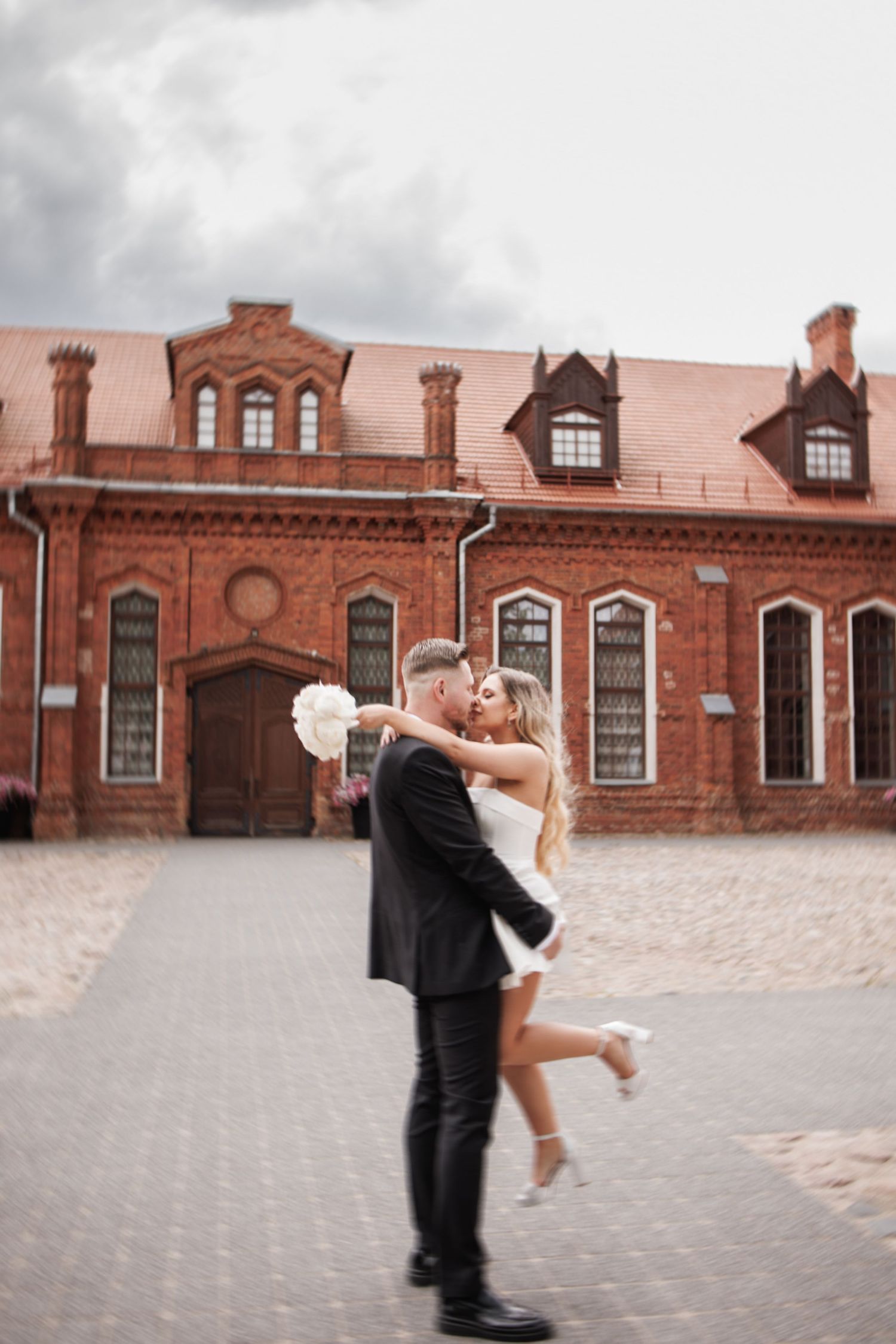 Romantic couple embraces in front of historic red brick building with ornate architecture and dormer windows.