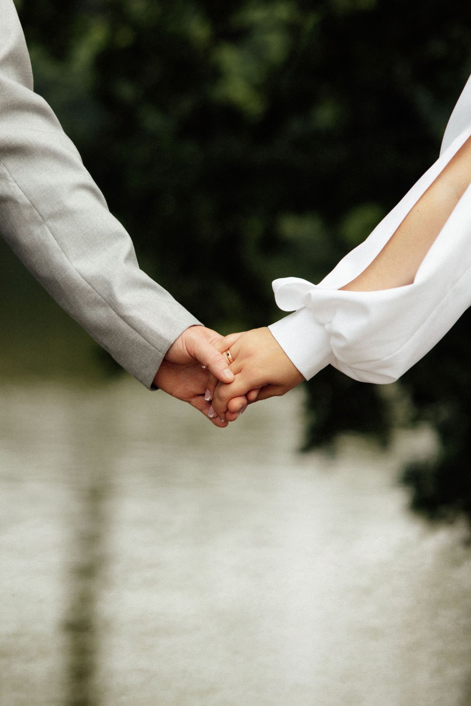 A bride and groom holding hands outdoors, showcasing a gold wedding ring, with a blurred green nature background.