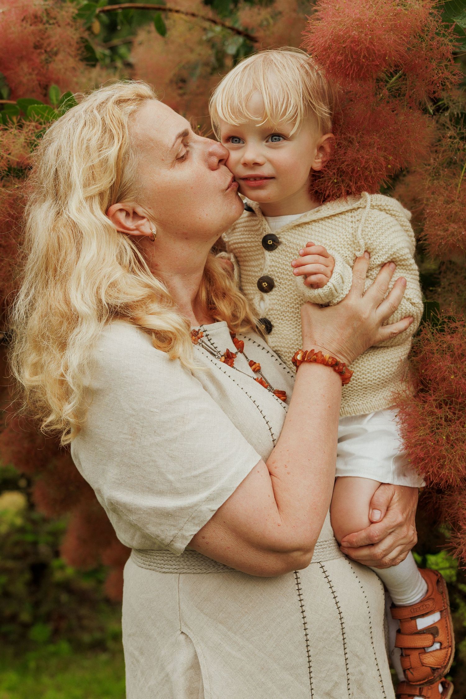 Blonde woman kissing a smiling toddler boy in a beige knit sweater outdoors near red flowering bushes.