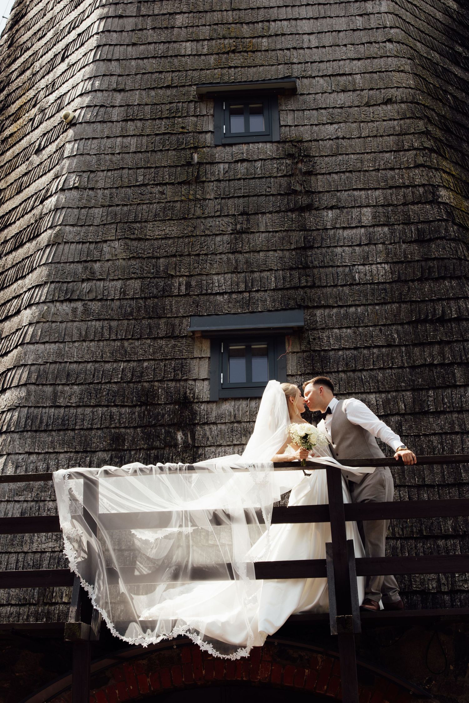 Bride and groom share a romantic kiss on a wooden balcony against a rustic stone building on their wedding day.