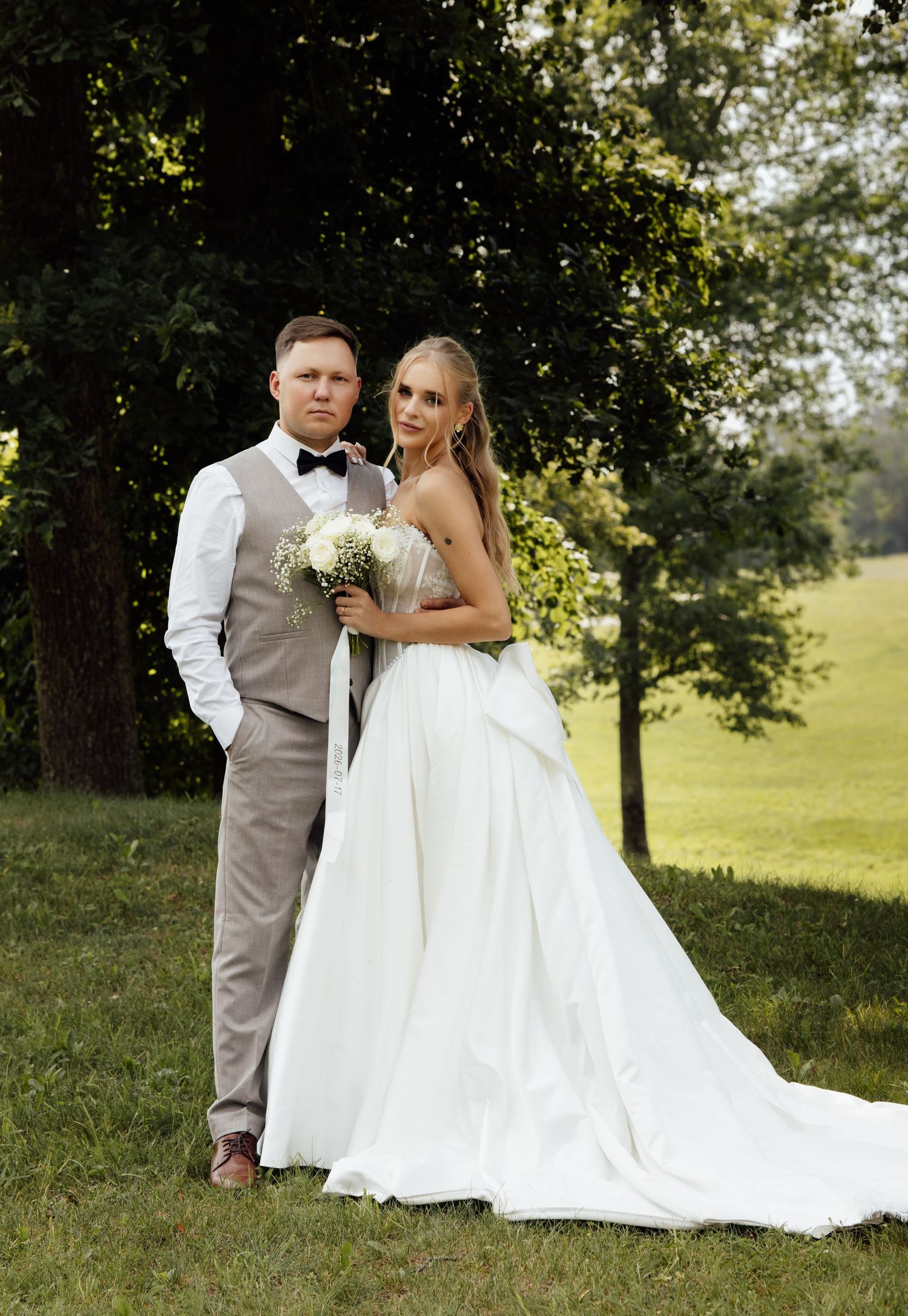 Bride holding bouquet and groom in gray vest posing together in a lush green outdoor setting.