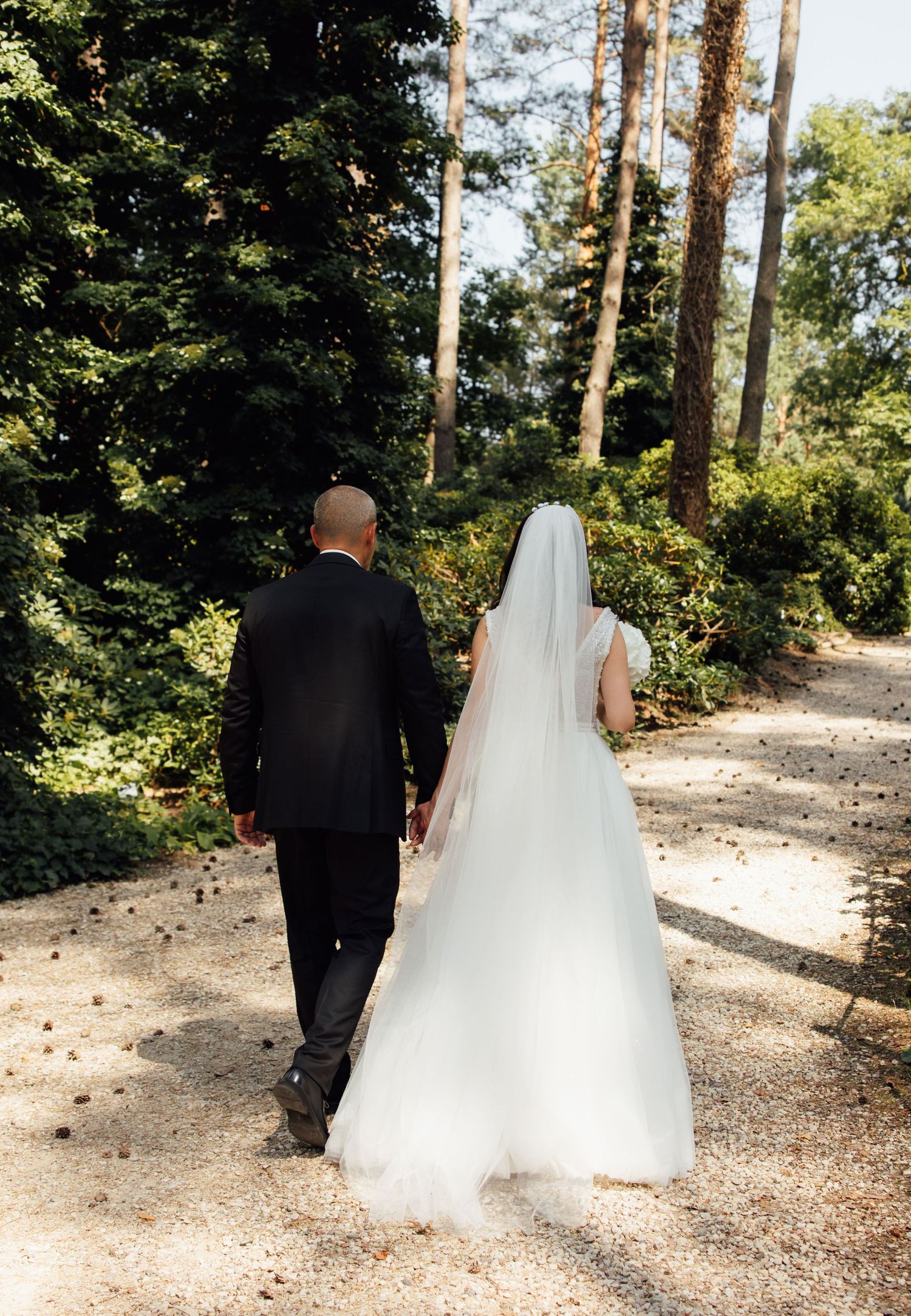 Bride and groom walking hand in hand down a sunlit forest path, bride wearing a white gown and veil.