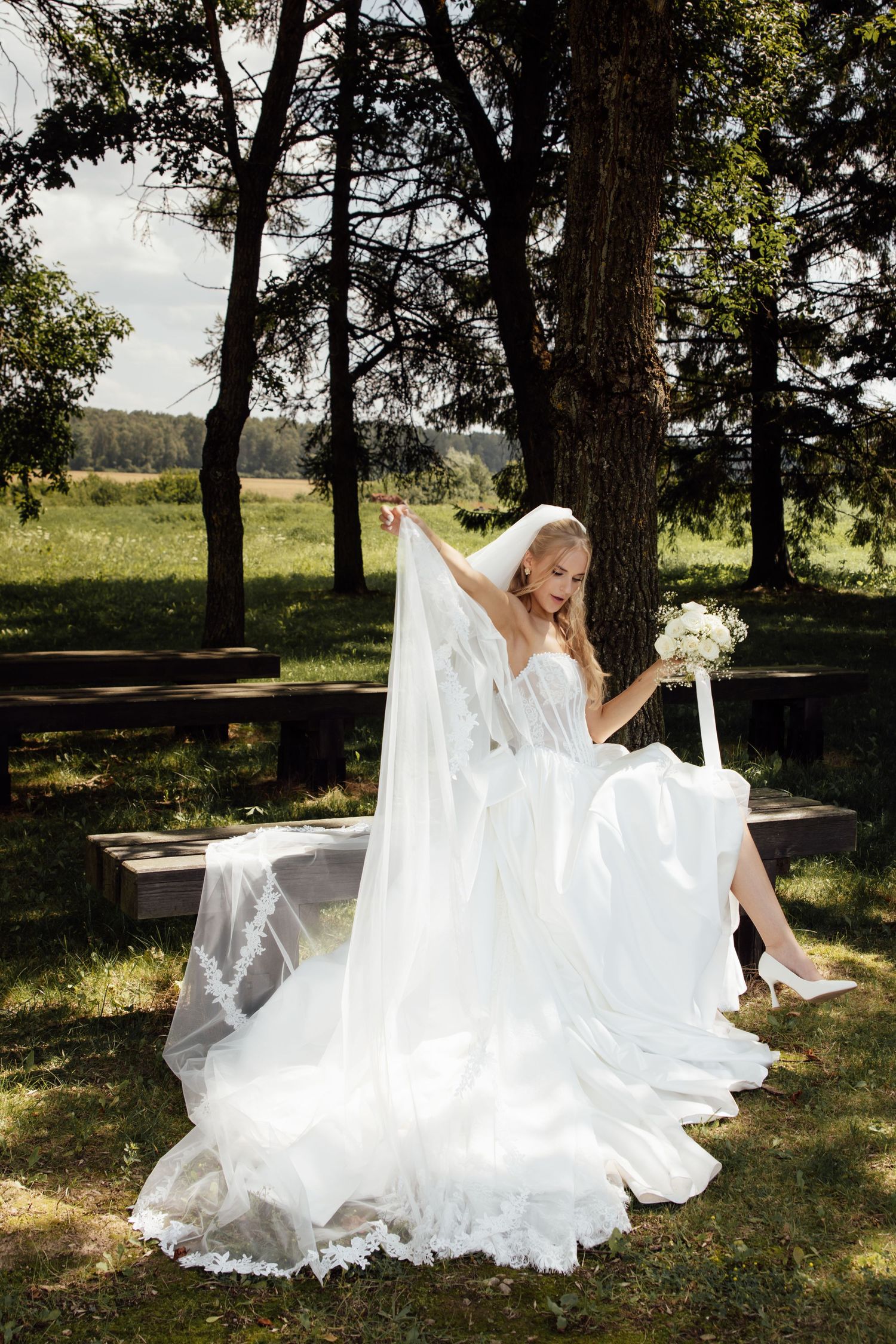 Elegant bride in white gown and veil sits outdoors under trees, holding bouquet, with flowing dress spread around her.
