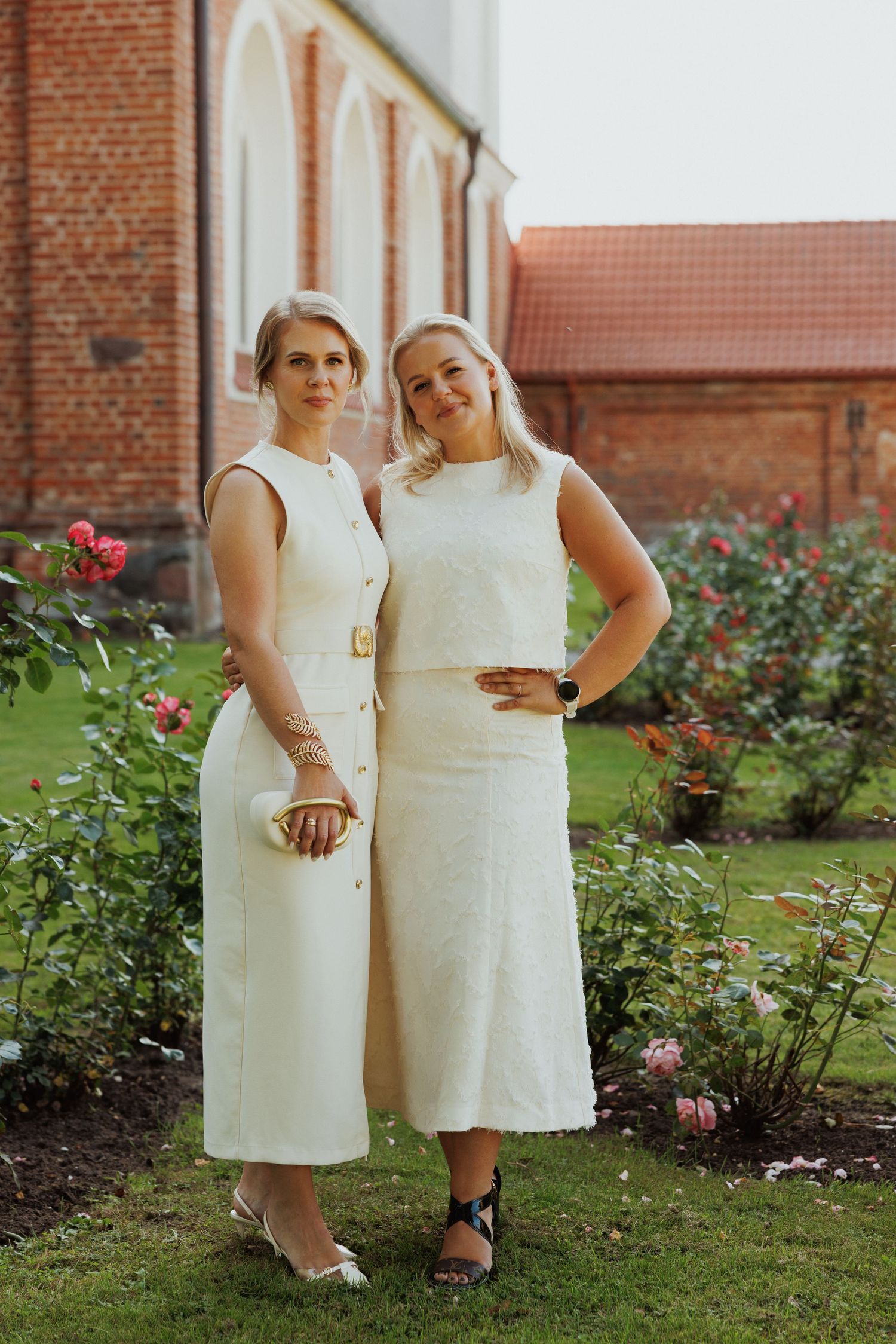Two people in elegant white outfits pose together in a garden with brick church backdrop.