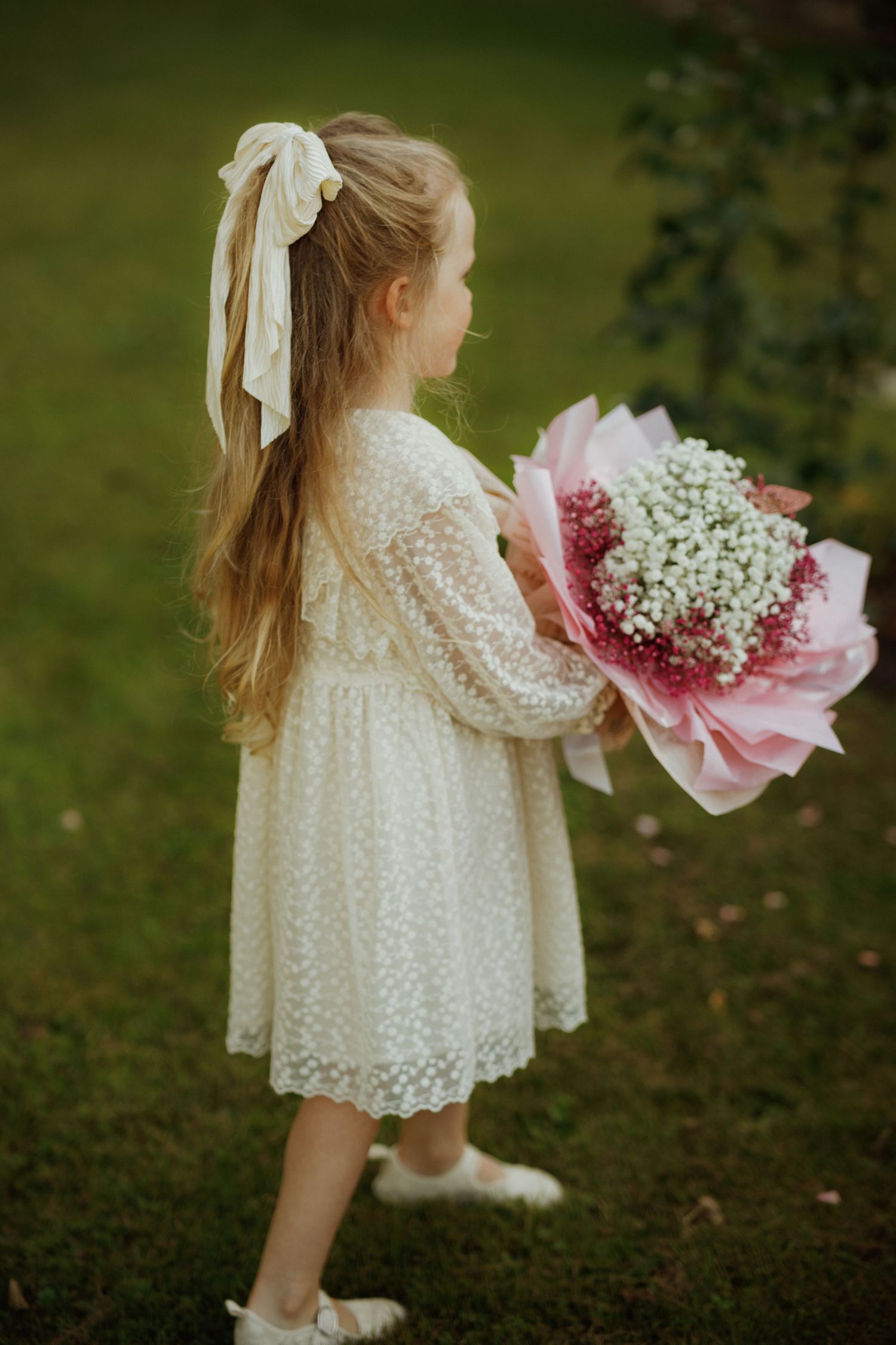 Little girl in white lace dress holding pink flowers while walking through a garden.