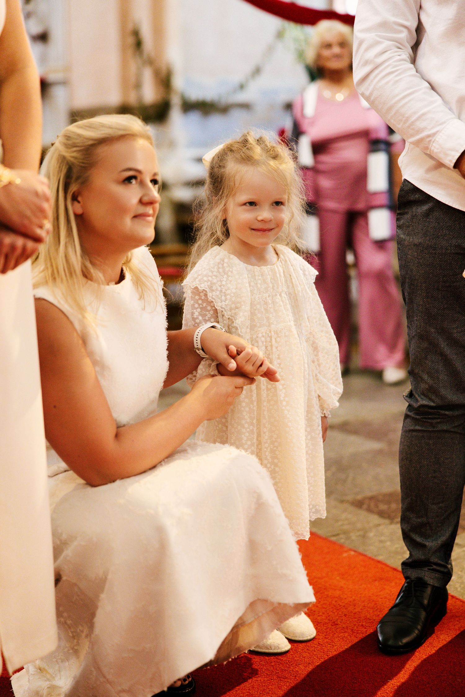 Young flower girls in white dresses sitting together on a red carpet during a wedding ceremony.