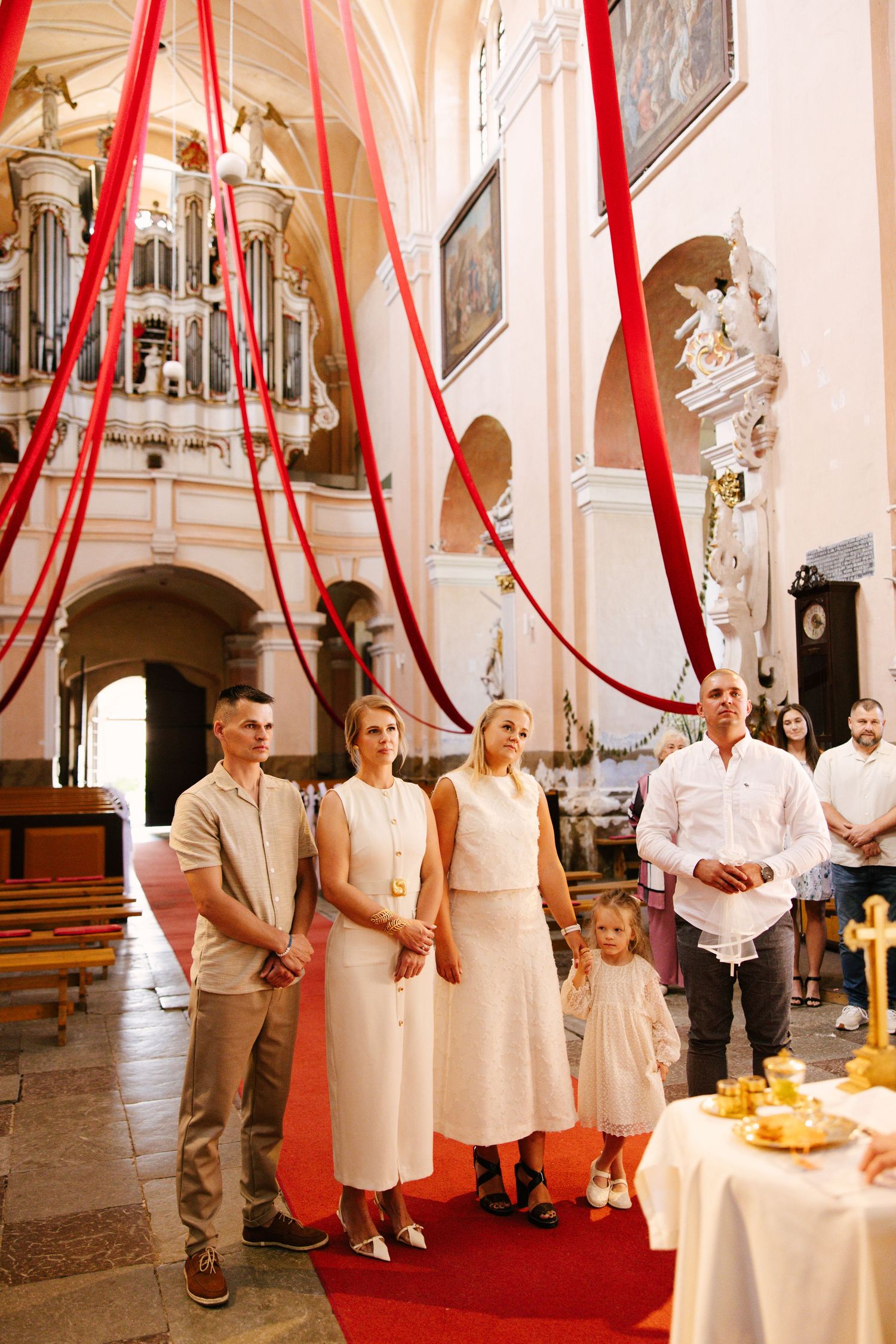 Interior of a church decorated with red ribbons and white decor during a ceremony with people gathered on a red carpet.