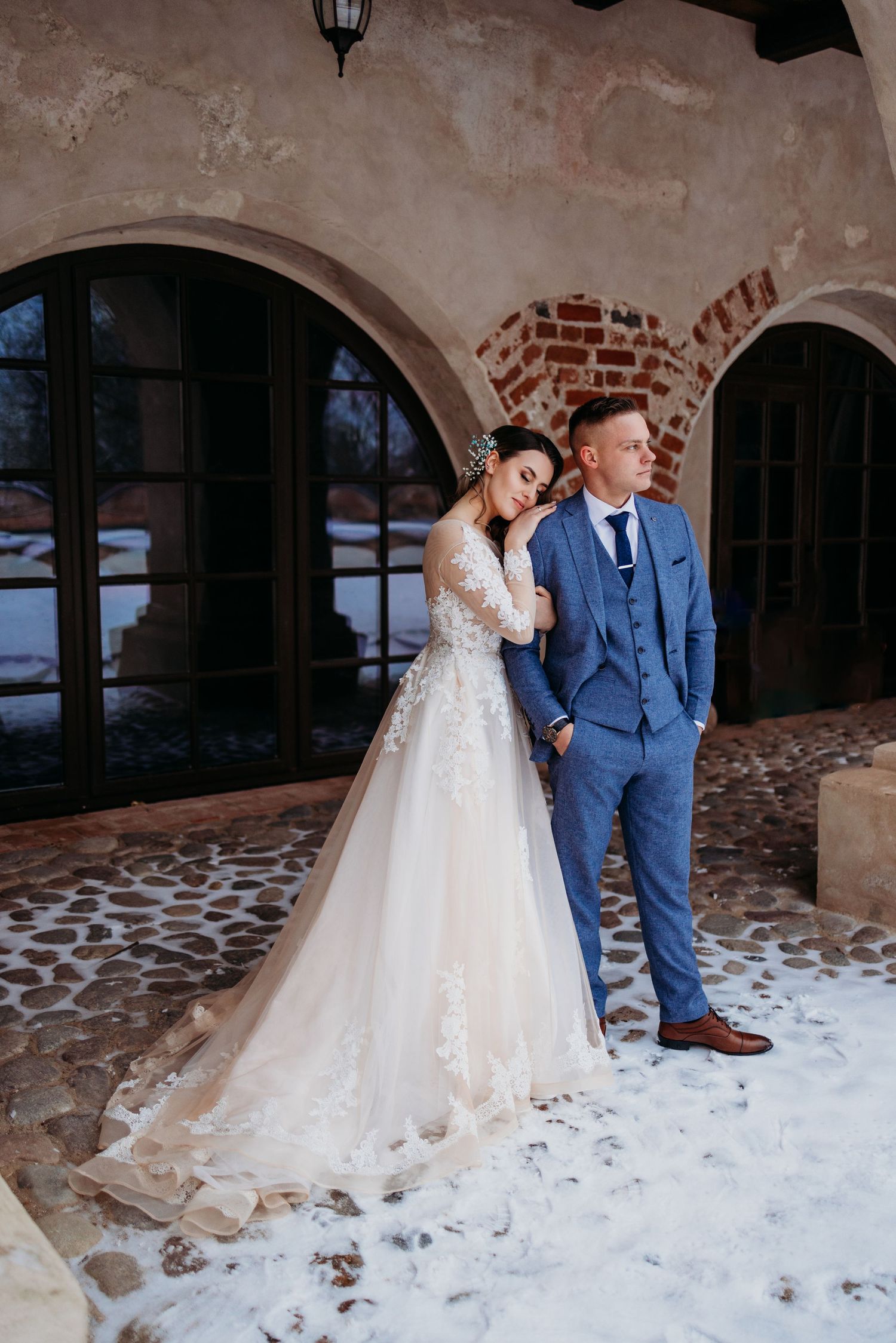 A romantic wedding couple poses against an arched stone wall in elegant attire.
