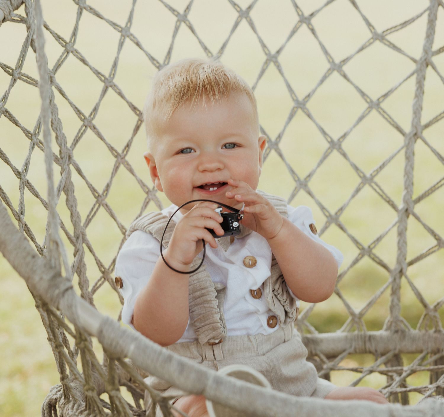 Baby in white outfit playing near a chain-link fence outdoors.