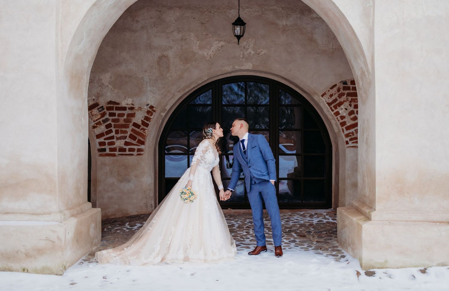 Wedding couple poses under stone archway in winter setting with exposed brick details.