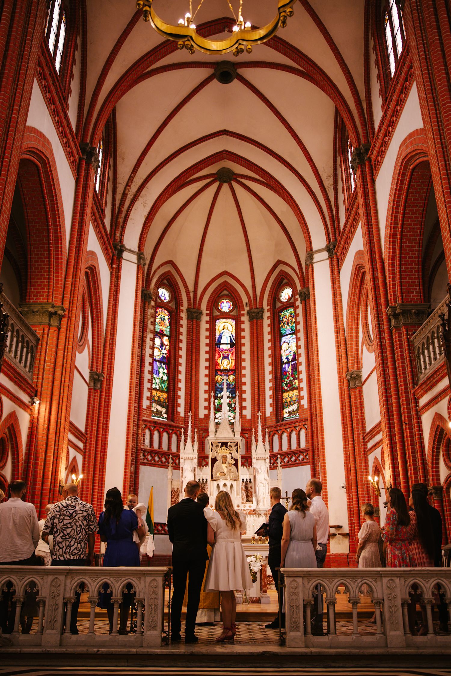 Interior view of a gothic cathedral with red brick arches, stained glass windows, and altar during a ceremony.