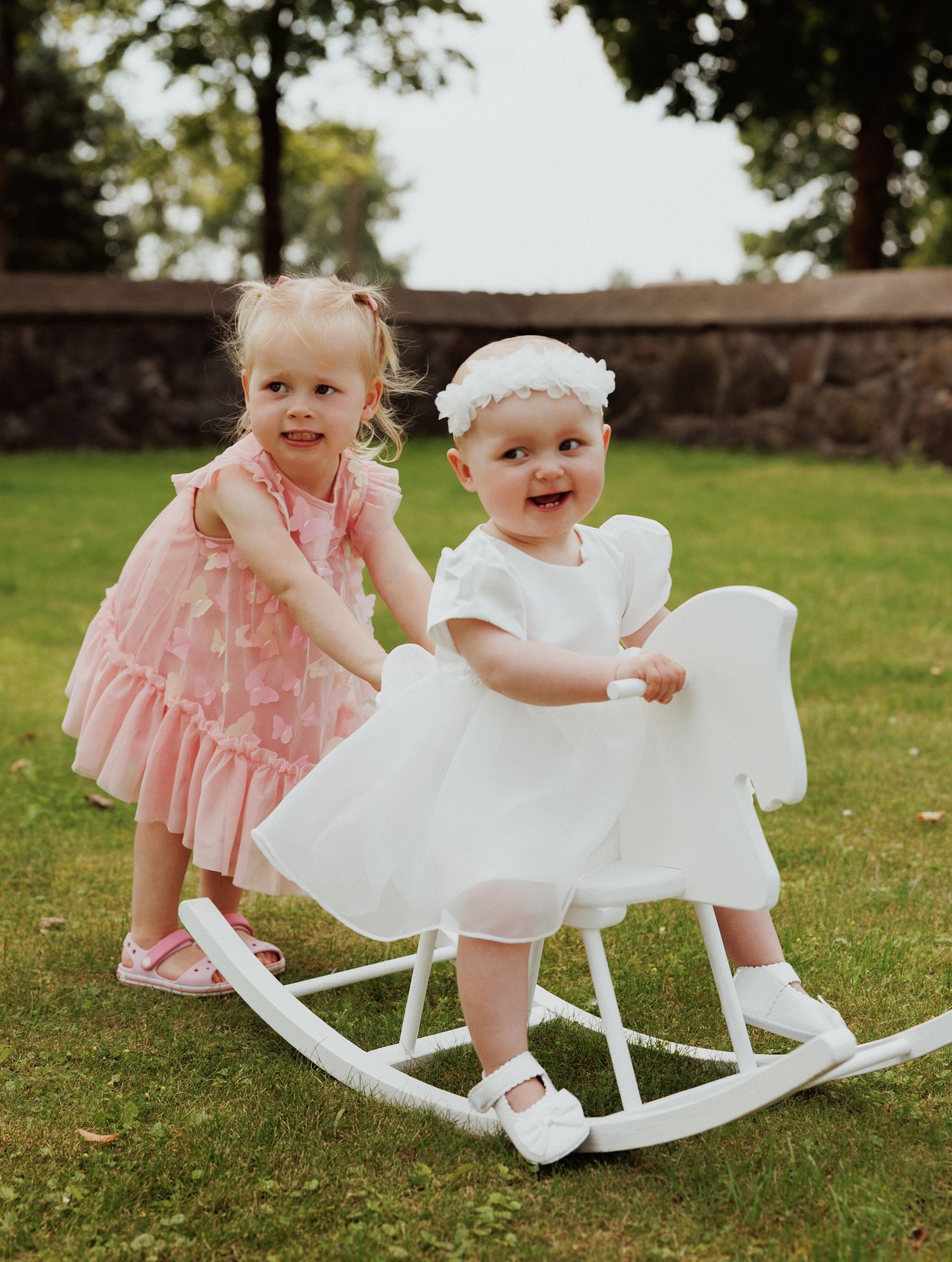 Small children playing on white rocking horses on a grassy lawn in pink and white dresses.