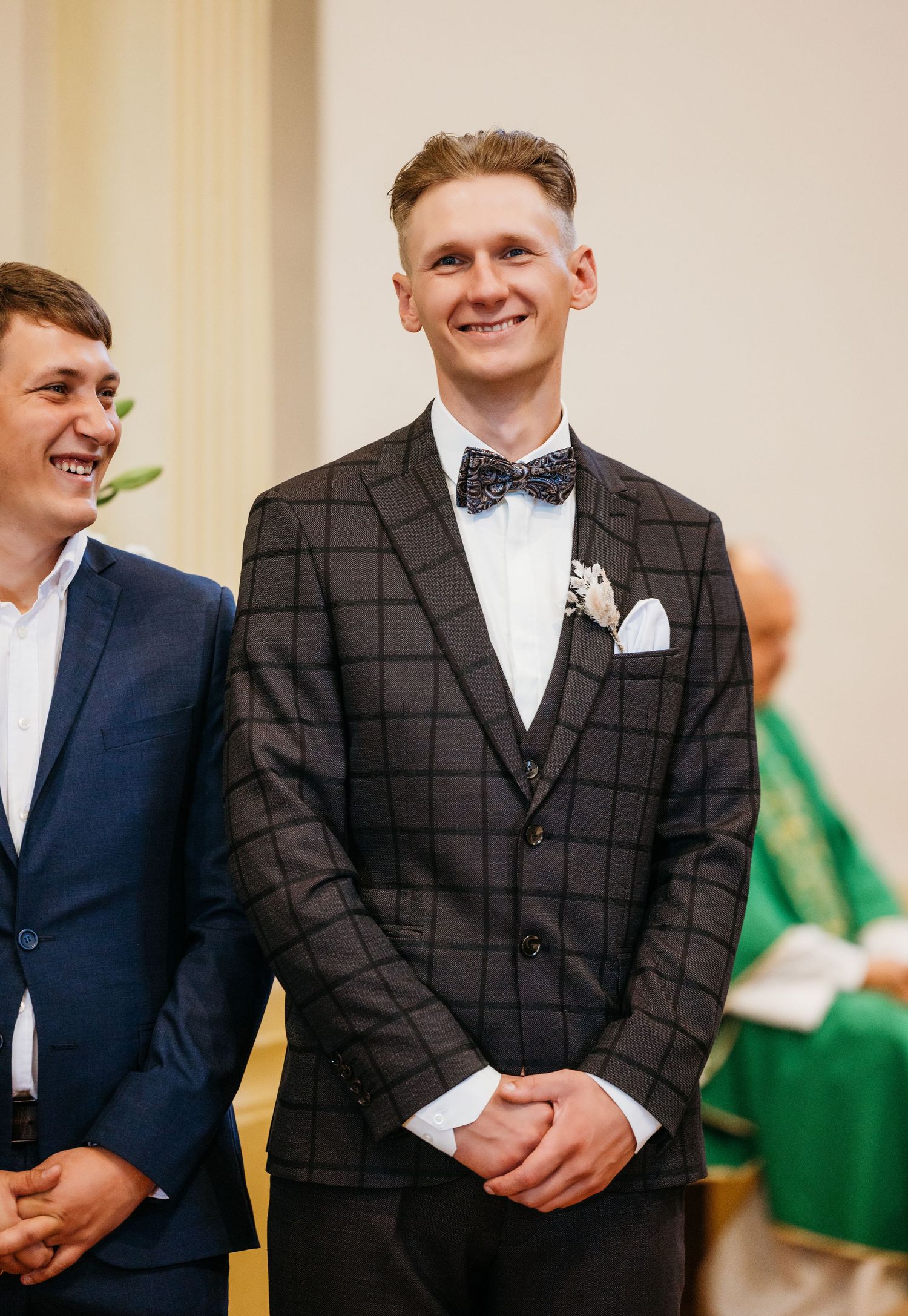 Groom in checkered suit and bowtie smiling at wedding ceremony.