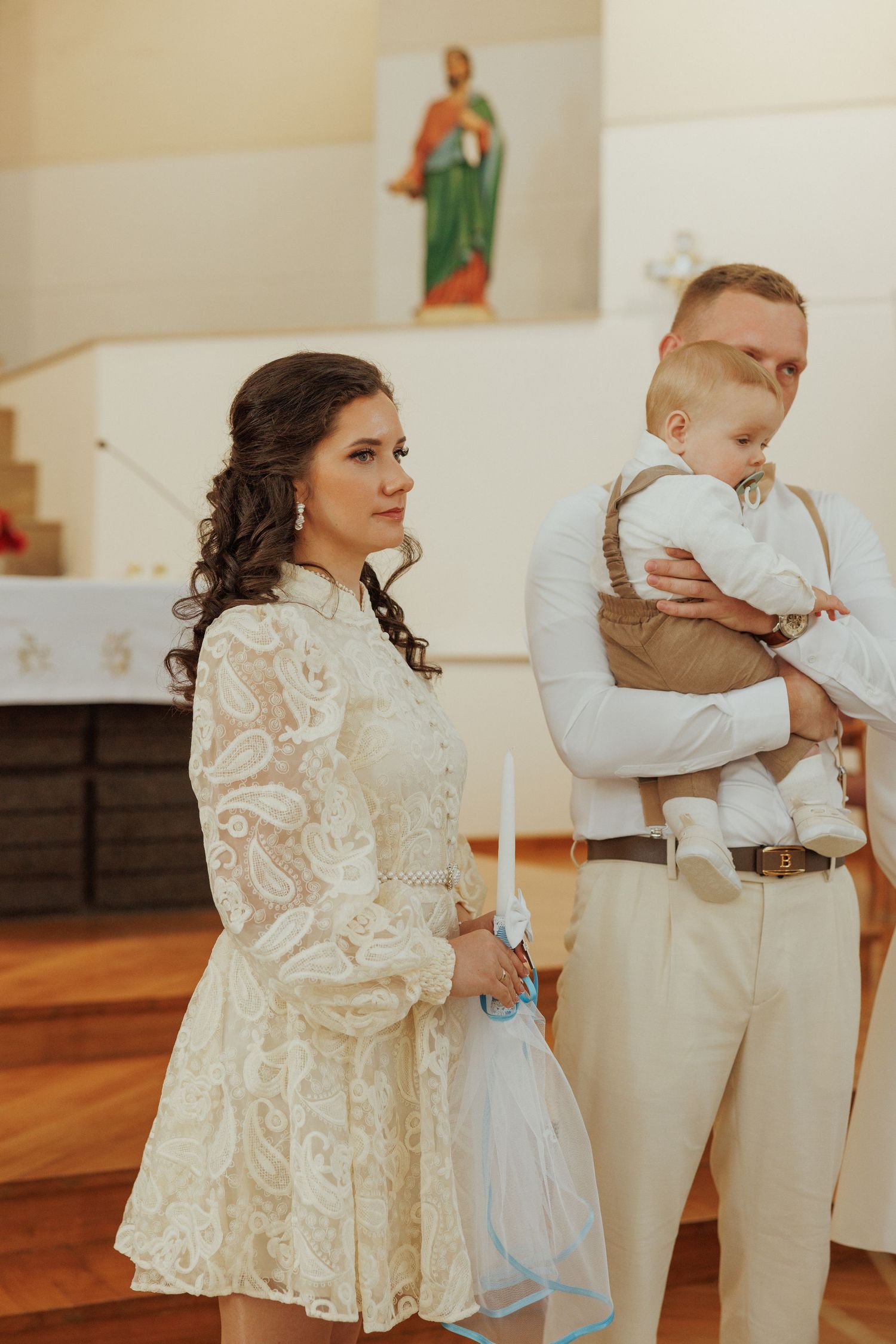 A baptism ceremony in a church with a couple in formal attire holding a baby while a religious statue overlooks.
