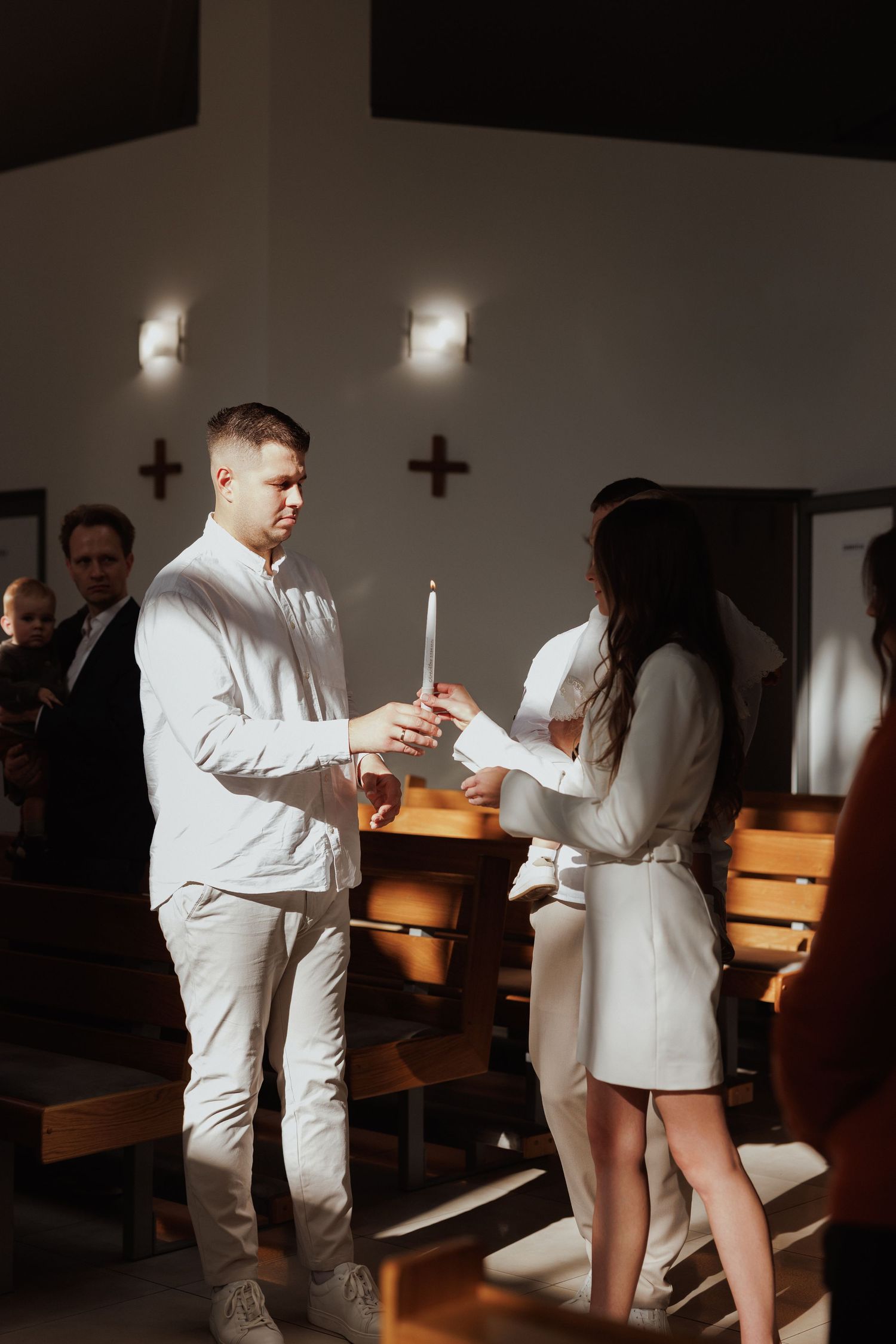 A couple in white outfits holds candles during an intimate church ceremony with wooden pews and crosses on the wall.