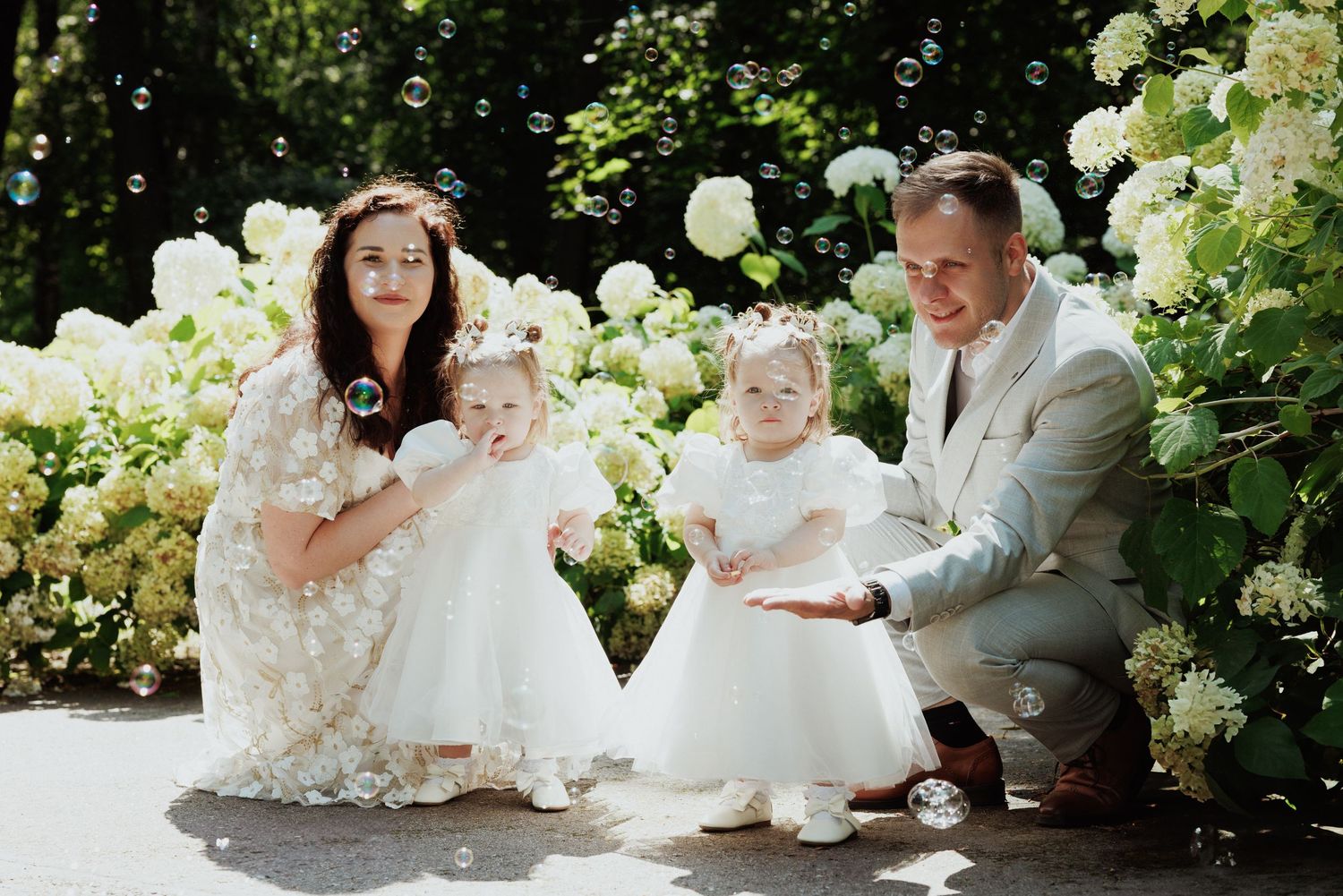 A family poses together in a lush garden setting wearing elegant white and cream colored formal attire.