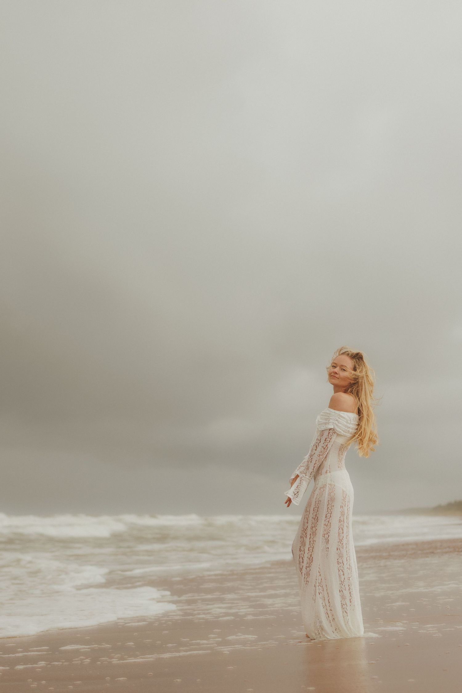 Blonde woman in white lace maxi dress standing on beach under overcast sky.