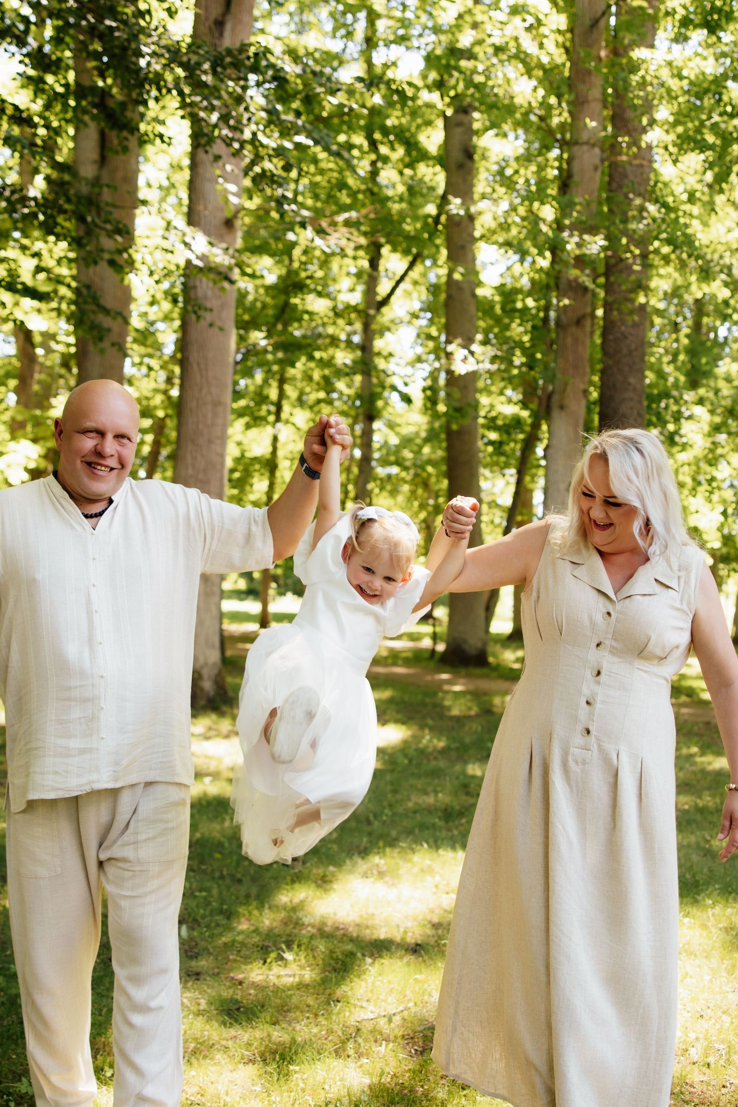 Happy family with bald father, blonde mother in beige dress swinging little girl in white dress outdoors in sunny park.