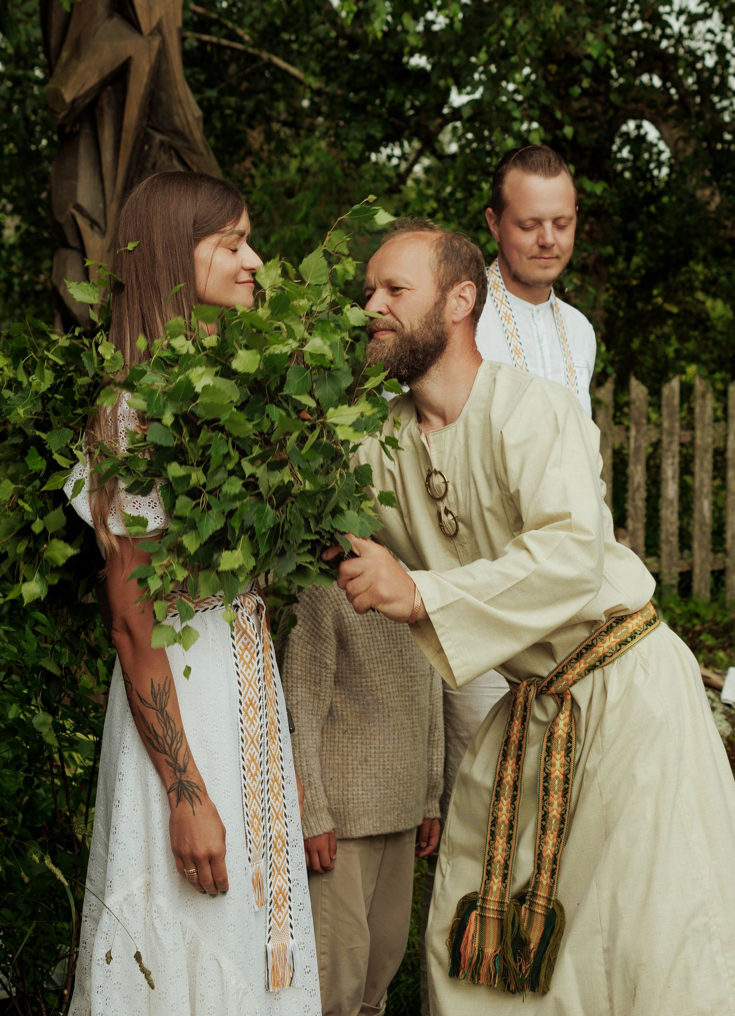 Three people in traditional folk attire with ivy garland celebrating outdoors in a garden setting.