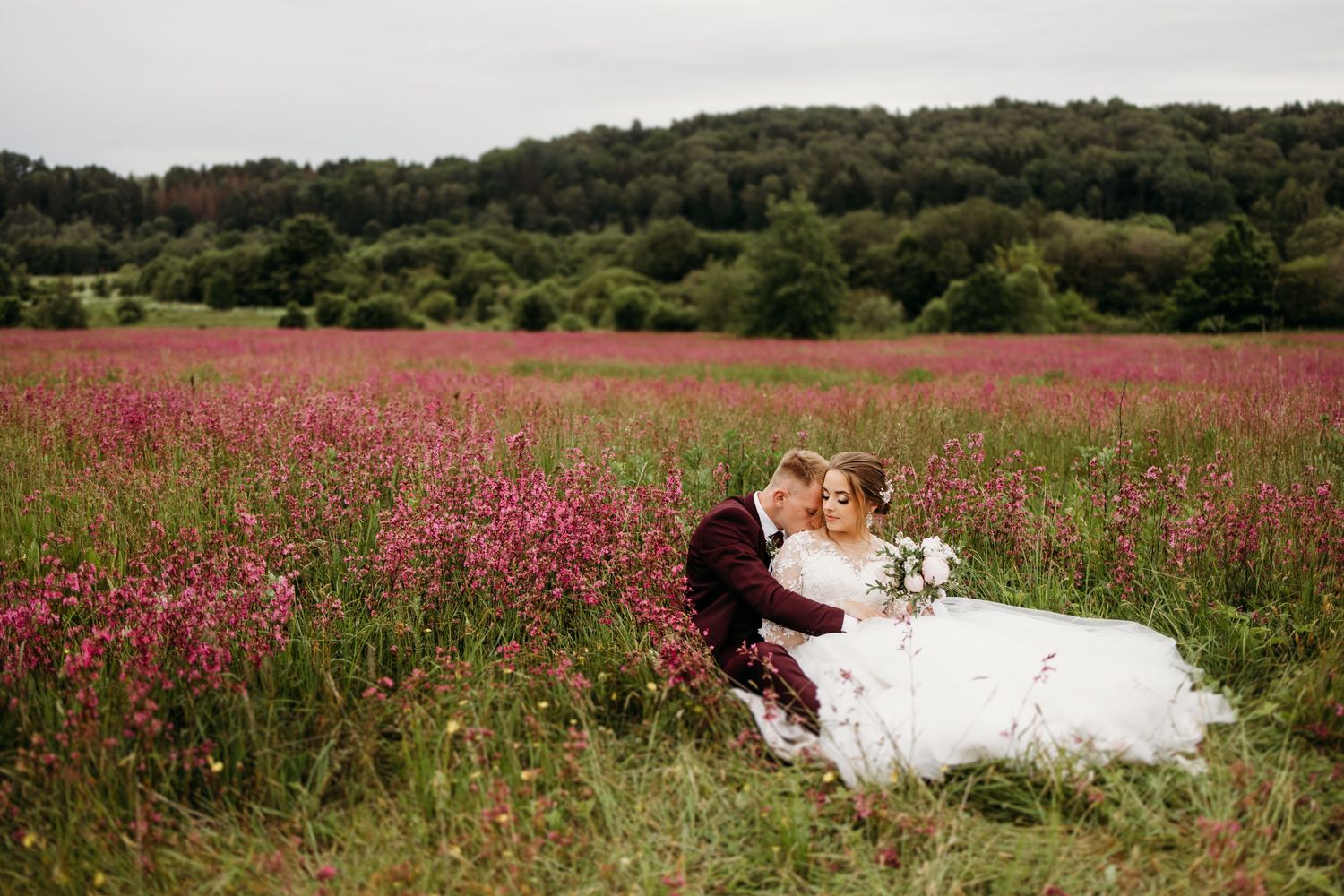 Wedding couple poses in scenic field with pink flowers and rolling hills in background.