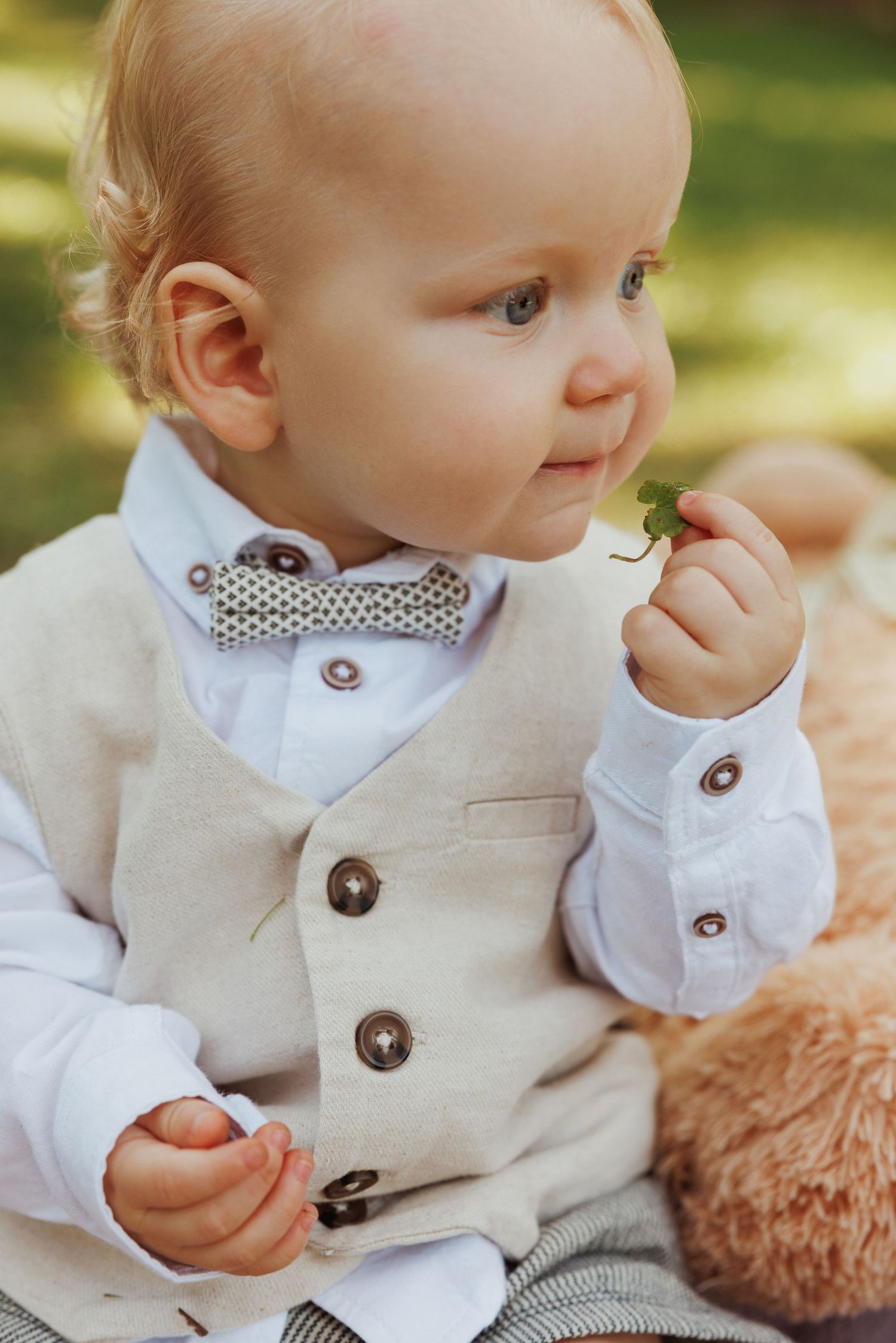 A baby in formal attire with polka dot bowtie and beige vest smiles while holding something small.