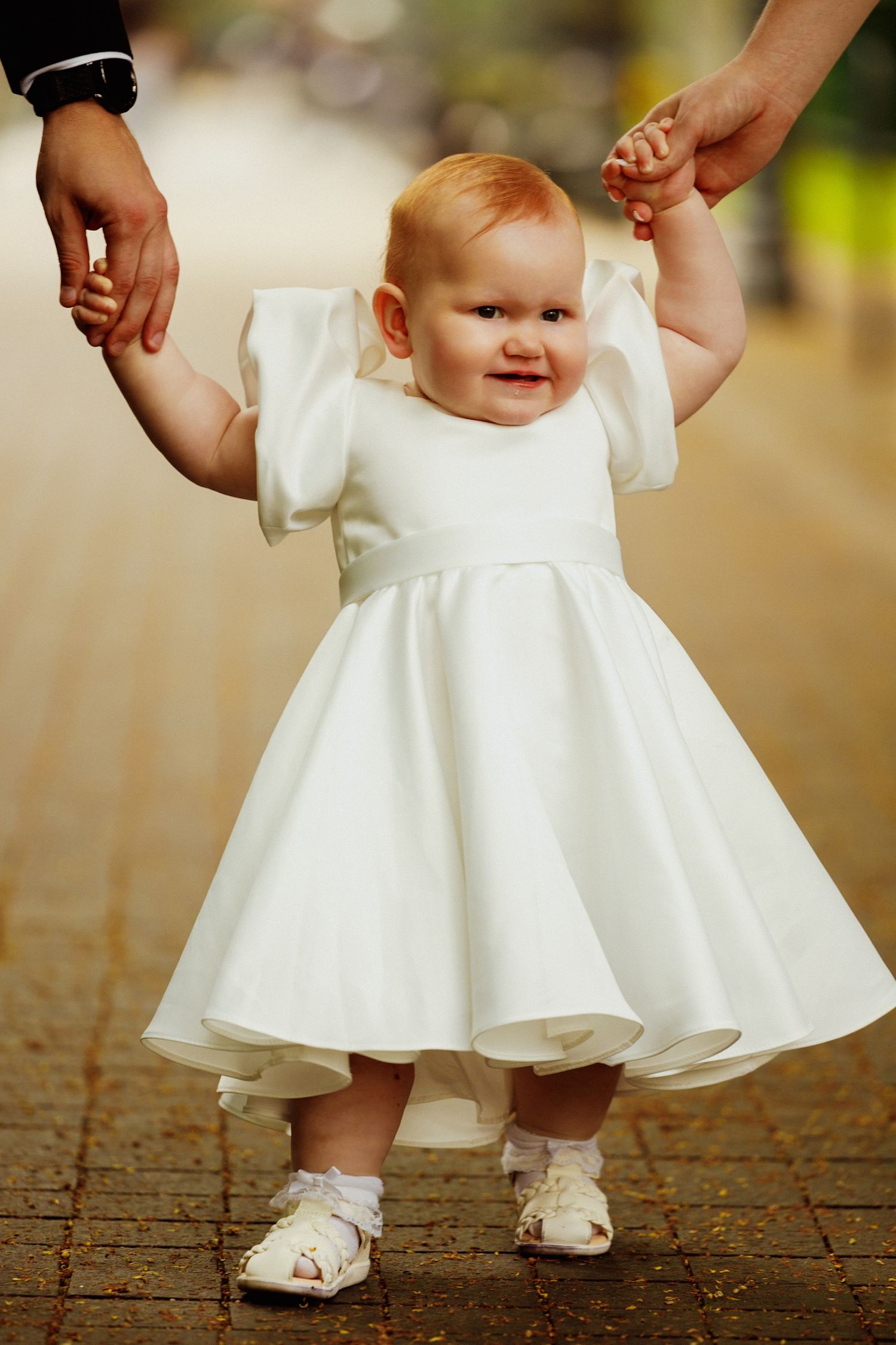 A small child in a white formal dress and sandals holds hands with adults while standing on brick pavement.