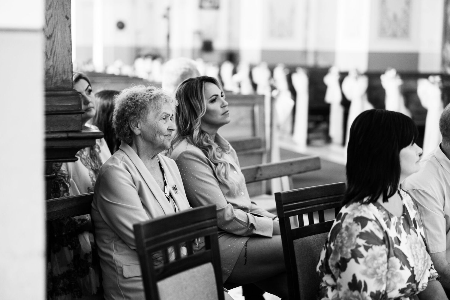 Two people sit together in wooden church pews while attending a service in black and white photography.