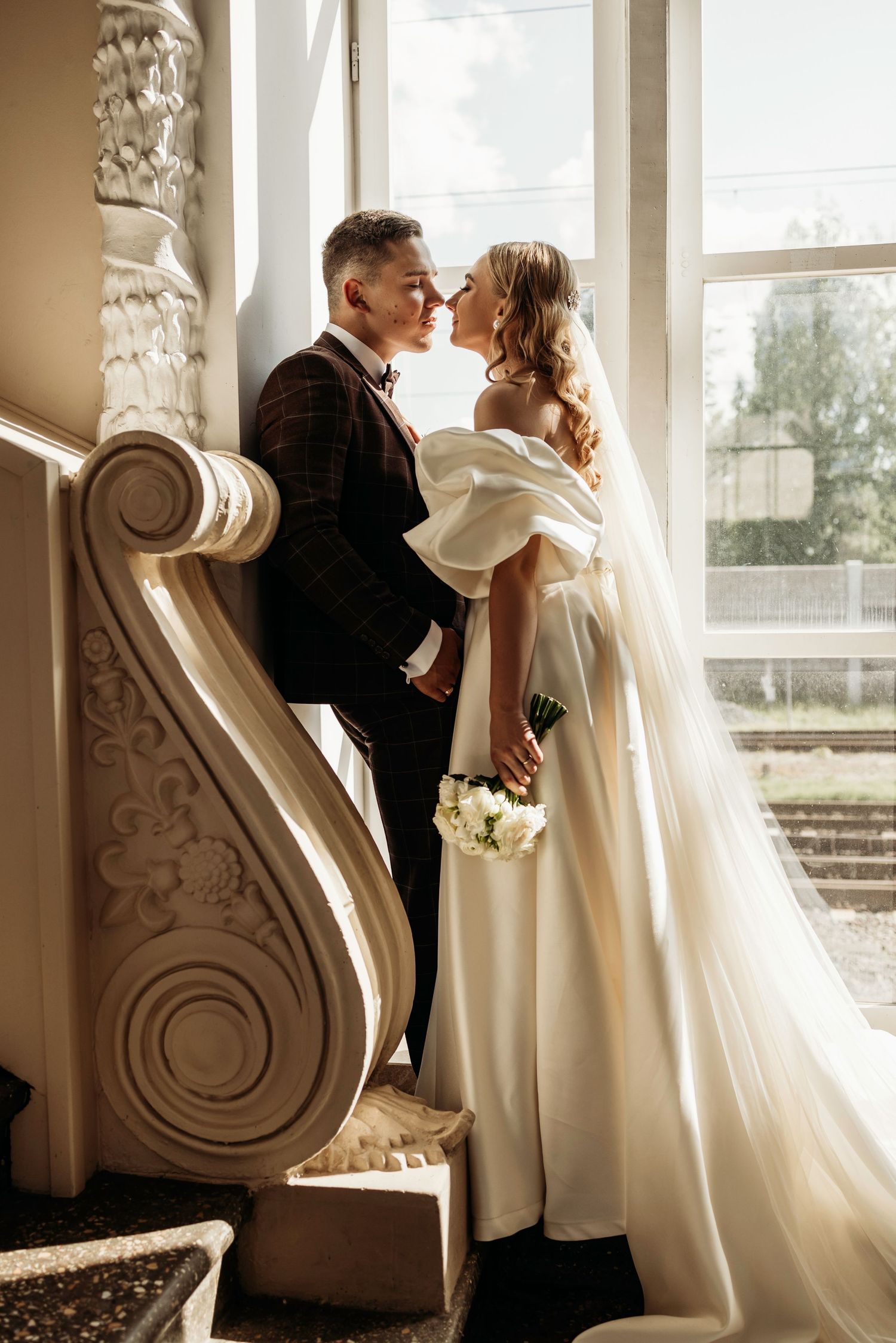 Romantic wedding couple in formal attire share intimate moments by ornate white columns in sunlit window setting.