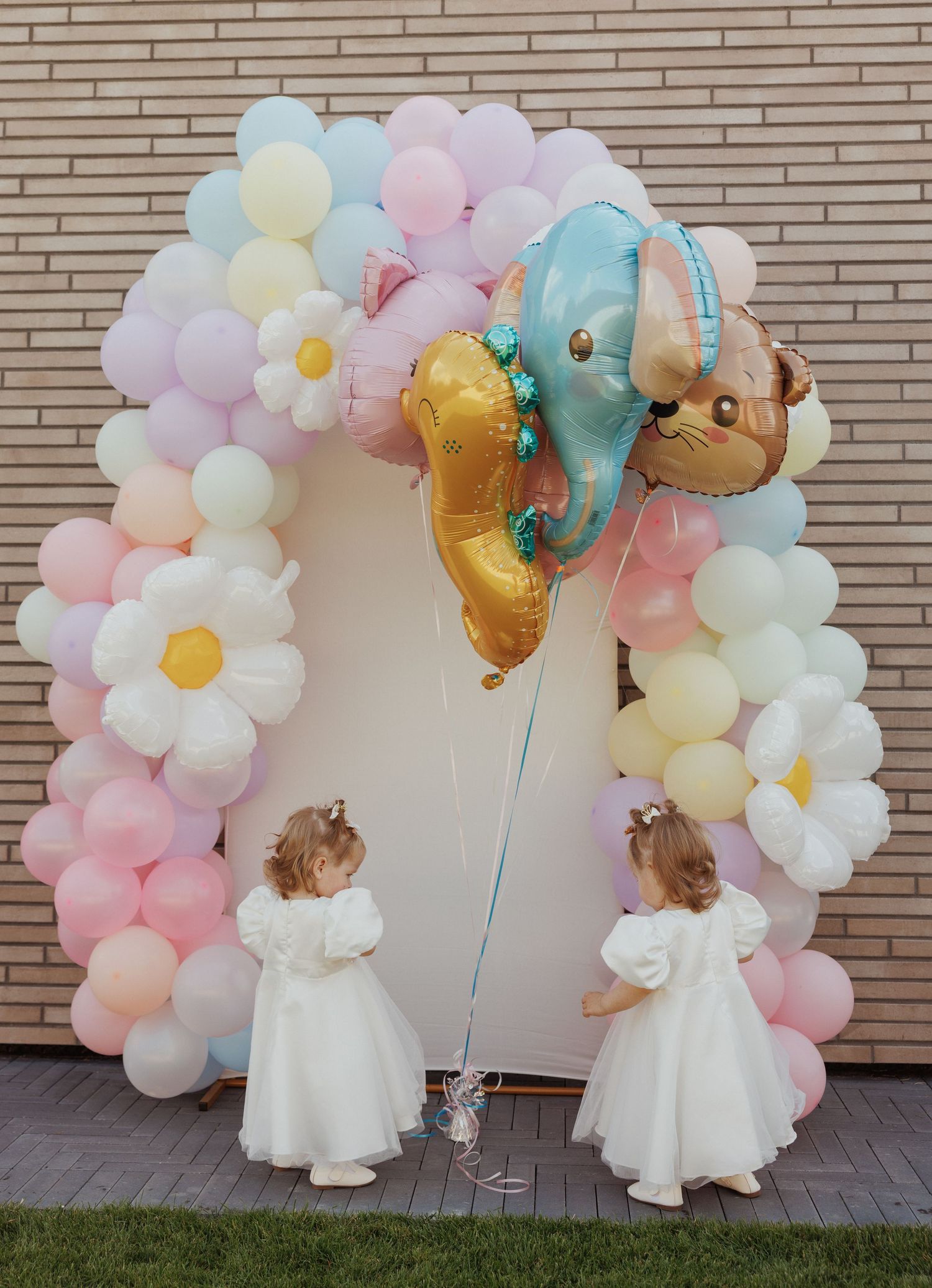 Whimsical balloon arch with pastel colors, daisies, and gold accents against brick wall.