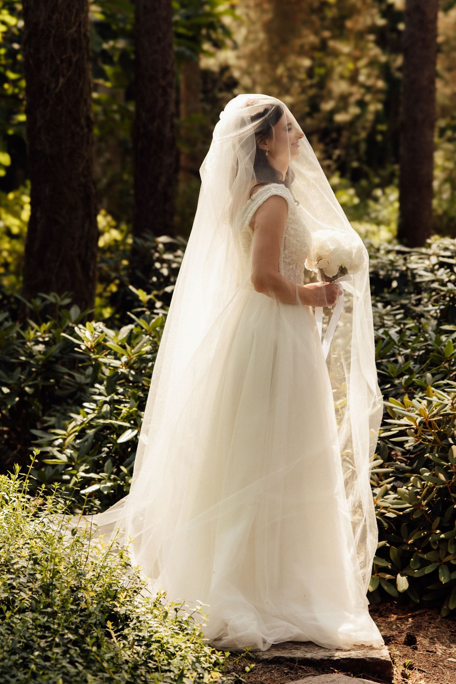 Bride in white tulle gown and long veil holds bouquet while standing in lush garden surrounded by green trees.