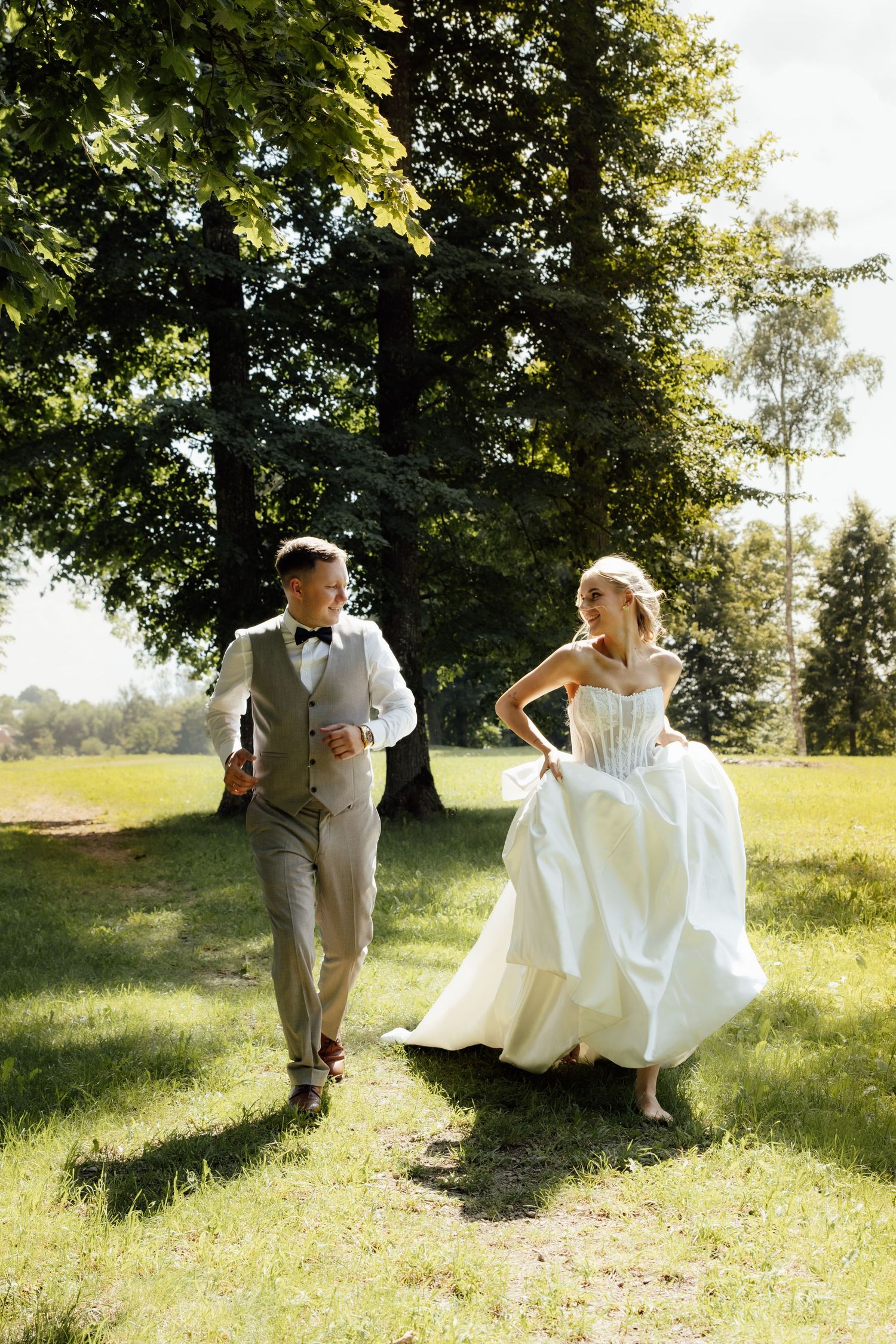 Joyful bride and groom run through sunny green park surrounded by tall trees on wedding day.
