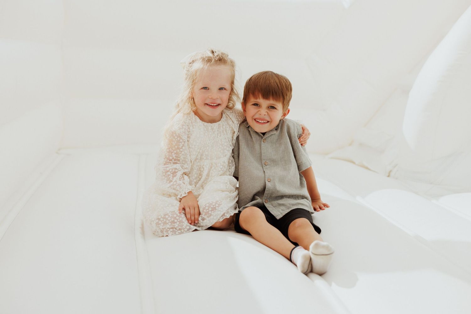 Two young children sit together and smile on a white backdrop during a photo session.