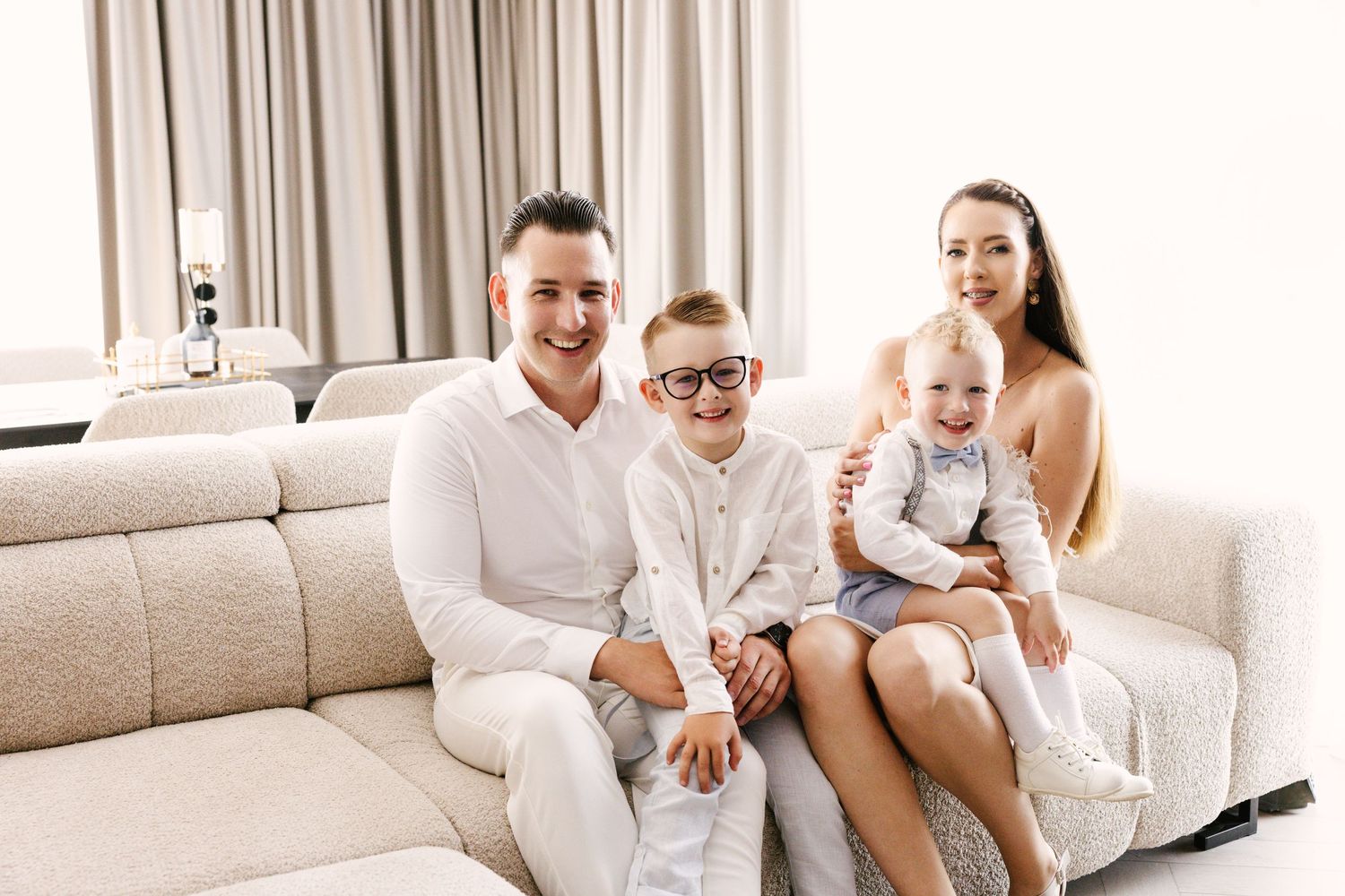 Happy family wearing white loungewear sits together on a beige couch in a bright living room.