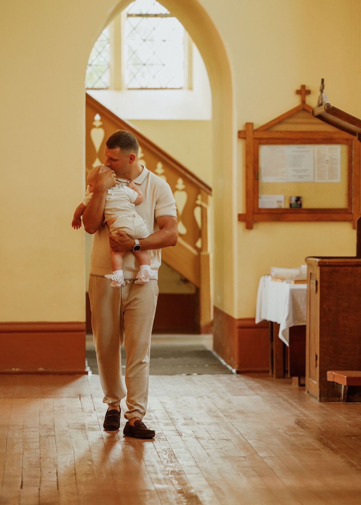 A parent in white clothing carries a sleeping baby through a sunlit church hallway with wooden pews.