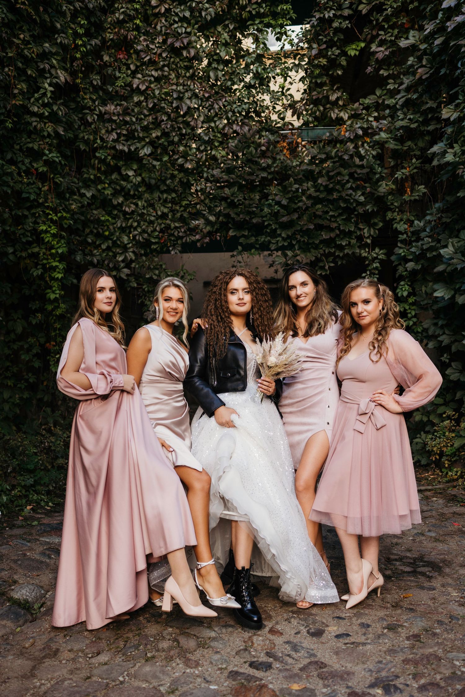 Bridesmaids in matching pink dresses pose playfully showing off their shoes against a leafy backdrop.