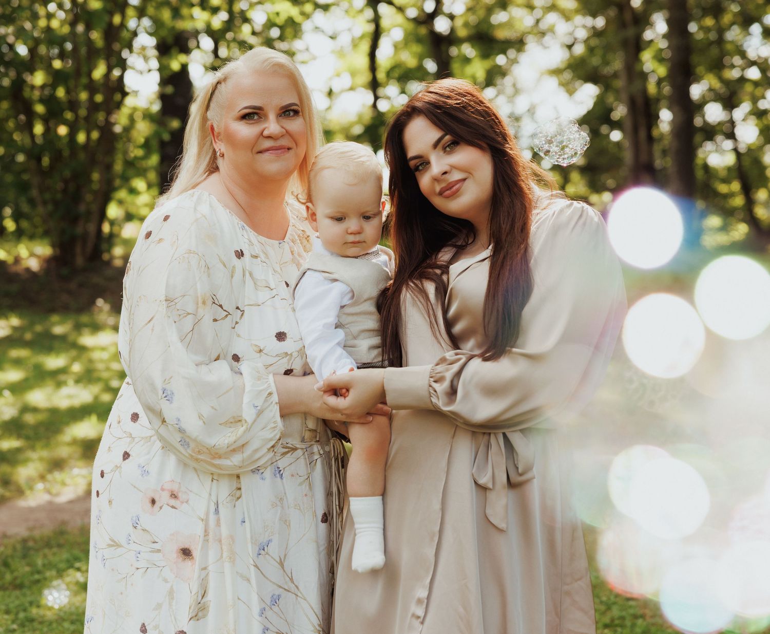 Three generations pose together outdoors in light-colored dresses on a sunny day with bokeh lighting effects.