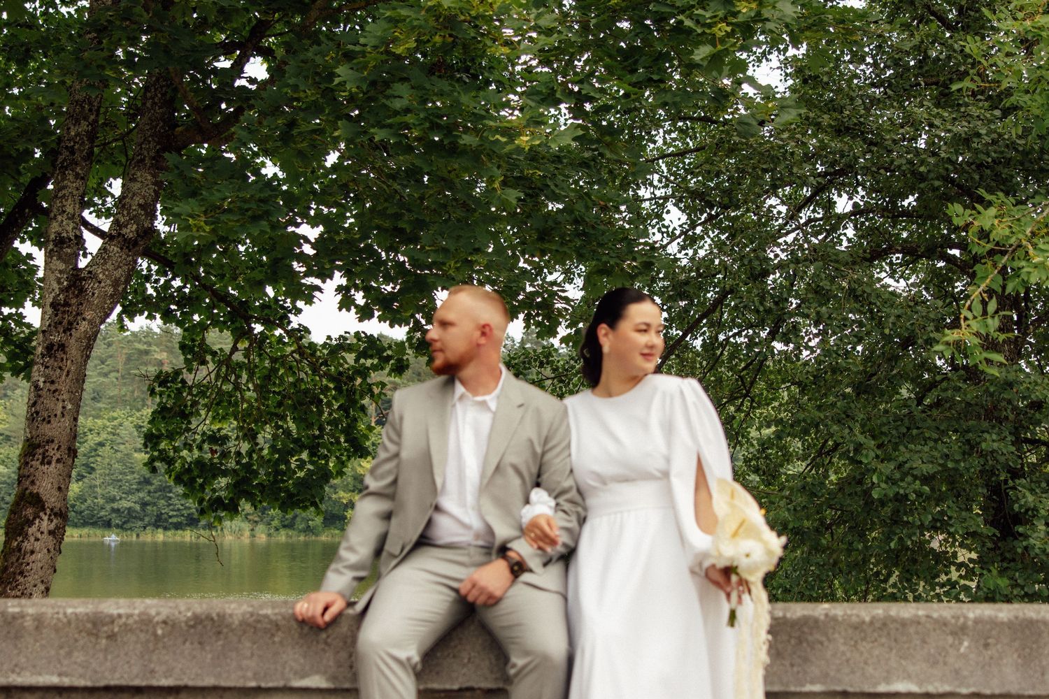 A couple in wedding attire sits on a stone wall surrounded by lush green trees near a lake.