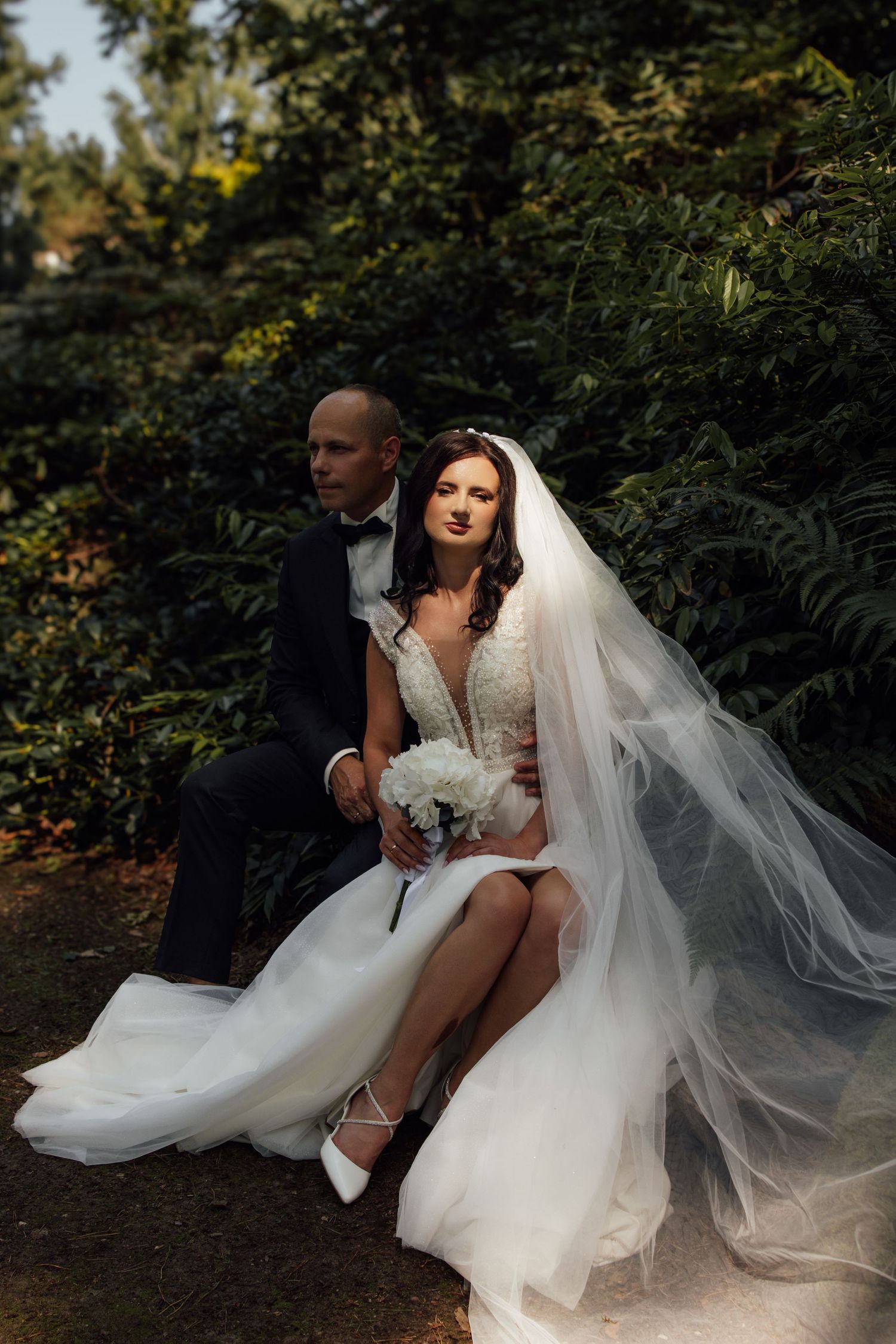 Bride and groom sitting together in lush garden, bride wearing white gown with veil, groom in black tuxedo.
